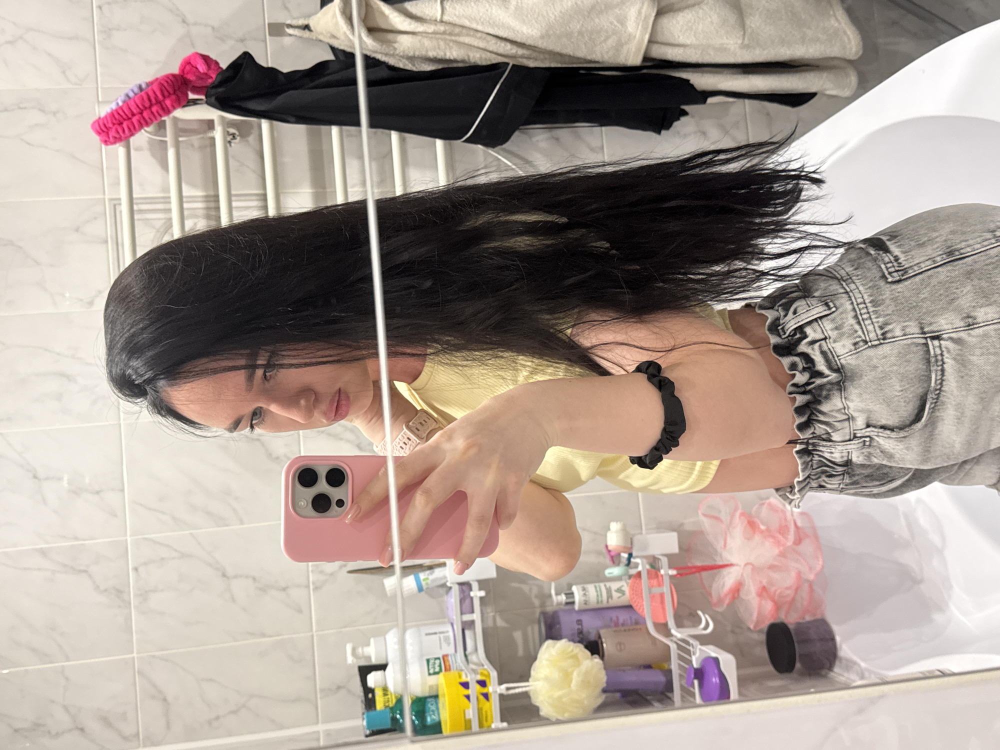 A young woman with long, dark hair is taking a mirror selfie in a bathroom. She is wearing a yellow crop top and gray denim shorts. She is holding a pink smartphone in her right hand, which is positioned to capture her reflection. To her left, a shelf is visible, stocked with various toiletries and bath products. A white towel rack with a pink bath accessory hanging from it is positioned behind her to the right. A white bathtub is visible in the lower portion of the image, reflecting the scene. The bathroom walls are tiled with a marble pattern. The overall setting suggests a private residential bathroom. The location in Novosibirsk, Russia cannot be determined from the visual information.