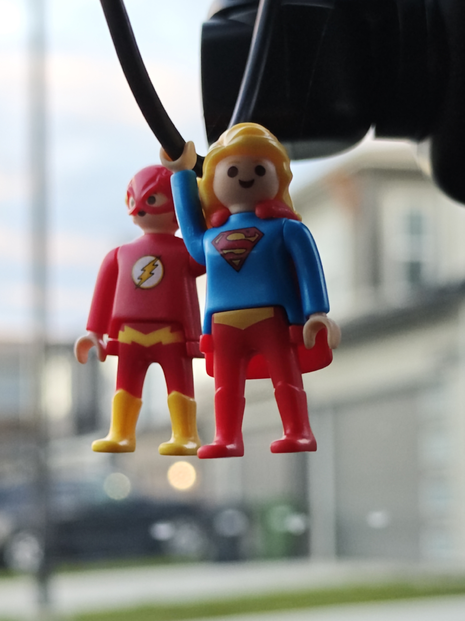 Two Playmobil figurines, one representing The Flash and the other Supergirl, are suspended by a black cord against a blurred urban background. The Flash figurine, on the left, wears a red suit with yellow boots and lightning bolt emblem. Supergirl, on the right, has blonde hair and a blue suit with the Superman emblem. Both figurines are positioned to suggest movement or flight. The background suggests a residential area with buildings and possibly a street, indicating this scene is likely captured outdoors in a city environment. The image is taken with a shallow depth of field, blurring the surroundings to emphasize the figurines. This photo was taken in Calgary, Canada.