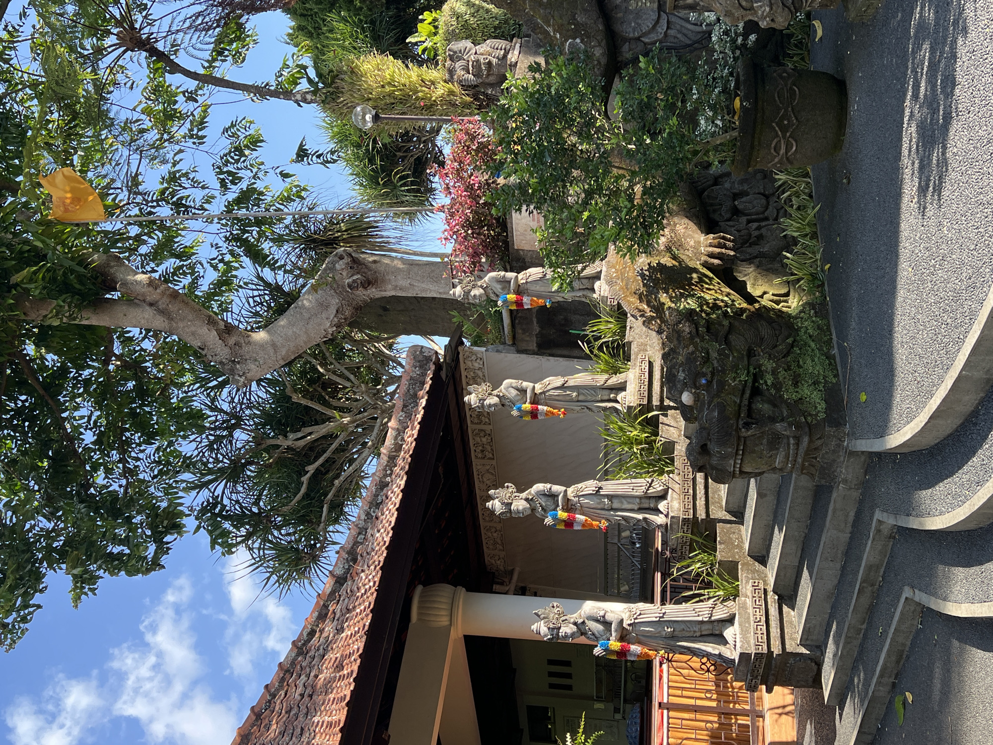 This is a low-angle, outdoor photograph taken in Pupuan, Indonesia. The scene features a traditional Balinese building with a red tiled roof on the left, leading to a covered walkway supported by columns. In front of the building and on tiered stone platforms are four stone statues of figures adorned with colorful garlands. The statues appear to be female, dressed in long robes, and wearing elaborate headdresses. A dragon-like stone carving is visible at the base of the stairs leading to the statues. Lush green foliage, including a large tree with a thick trunk, surrounds the statues and the building. A bright yellow flag is flying from a pole among the trees. The ground is paved with dark grey gravel and light grey concrete paths. The sky is blue with scattered white clouds.