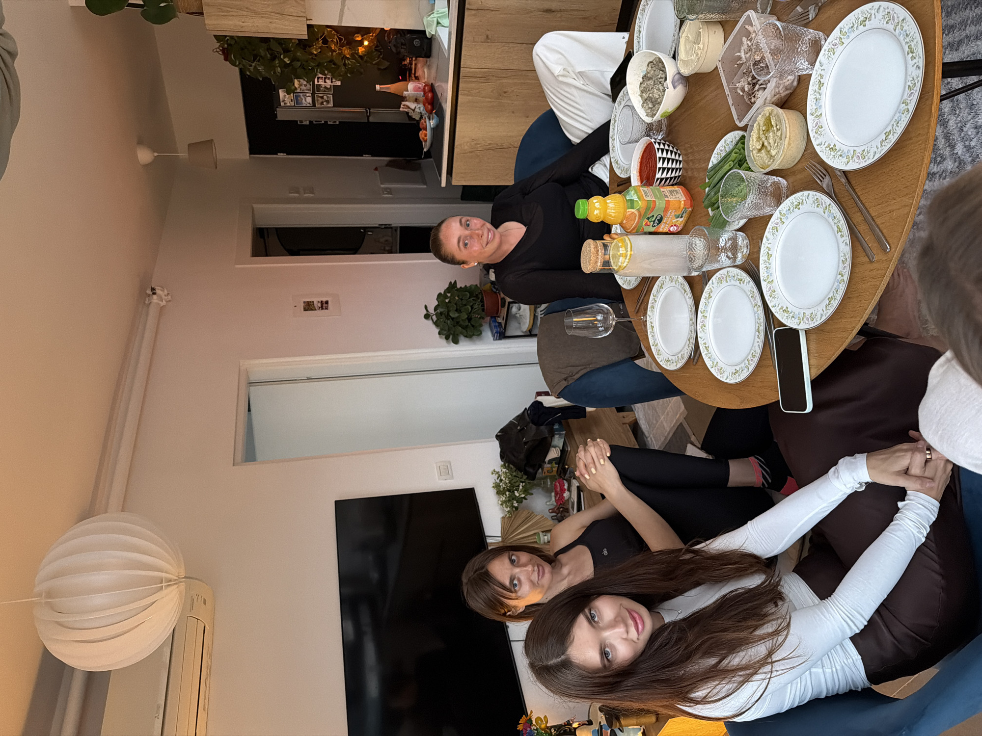 The image captures a casual gathering of three women around a round wooden dining table laden with various food items, likely in an apartment in Tel-Aviv, Israel. The table features plates, bowls, containers filled with dips and salads, and bottles of juice. Utensils are placed next to the plates. Two women are seated close together in the foreground, facing the camera with smiles. One is wearing a white top, and the other a black tank top. The third woman sits opposite them at the table, also facing the camera and wearing a black long-sleeve top. The apartment setting includes a television on the wall, indoor plants, and a decorative paper lantern hanging from the ceiling. The ambiance suggests a relaxed and informal meal or social event.