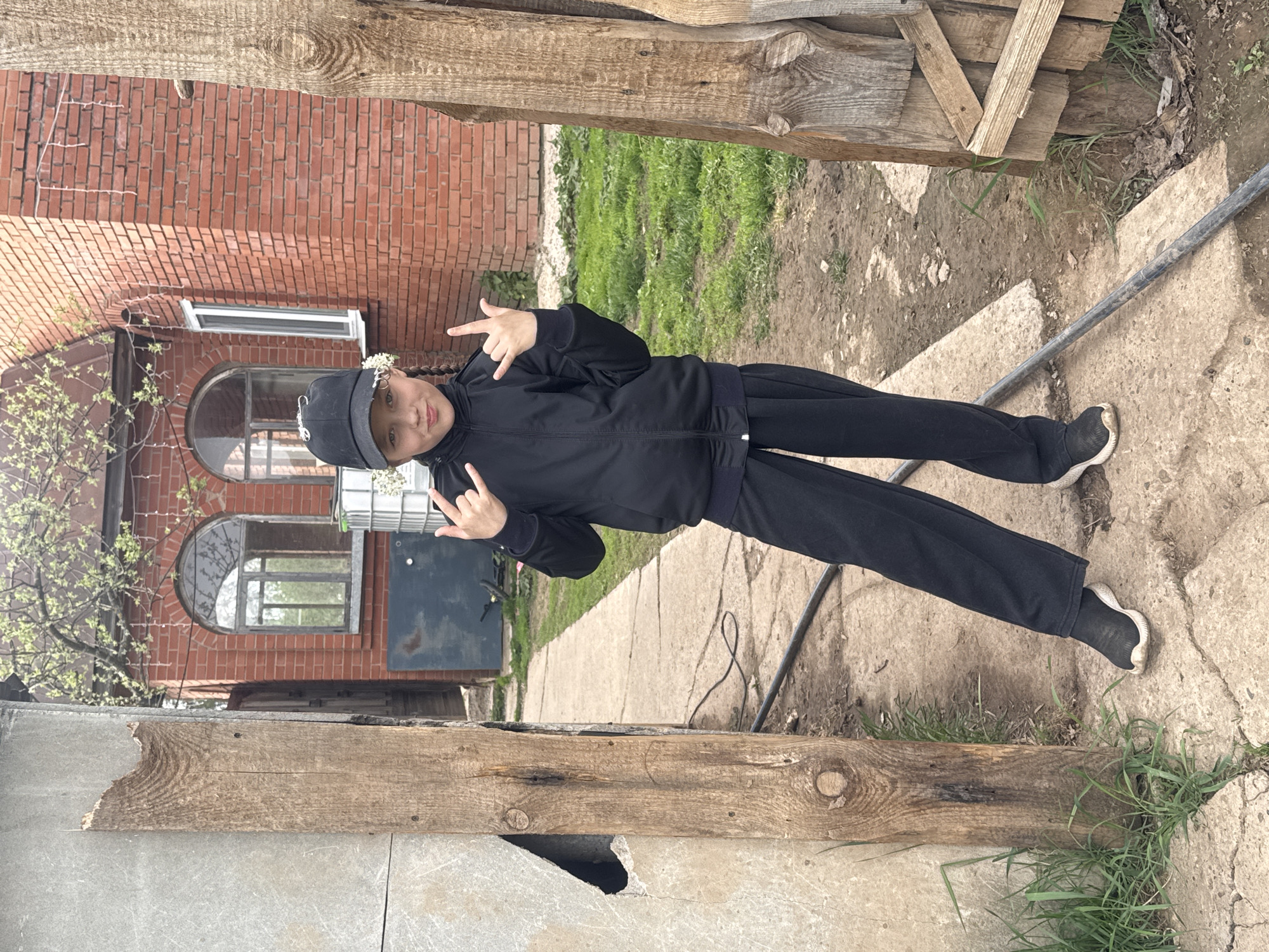 The image shows a girl in a black tracksuit standing outside. She is wearing a black cap and making the "rock on" hand gesture with both hands. A brick building with arched windows and budding trees is visible in the background. A concrete walkway and grassy patches are on the ground. Wooden planks frame the top and bottom of the image. The location is likely Rusalevka, Russia, based on the user's request.