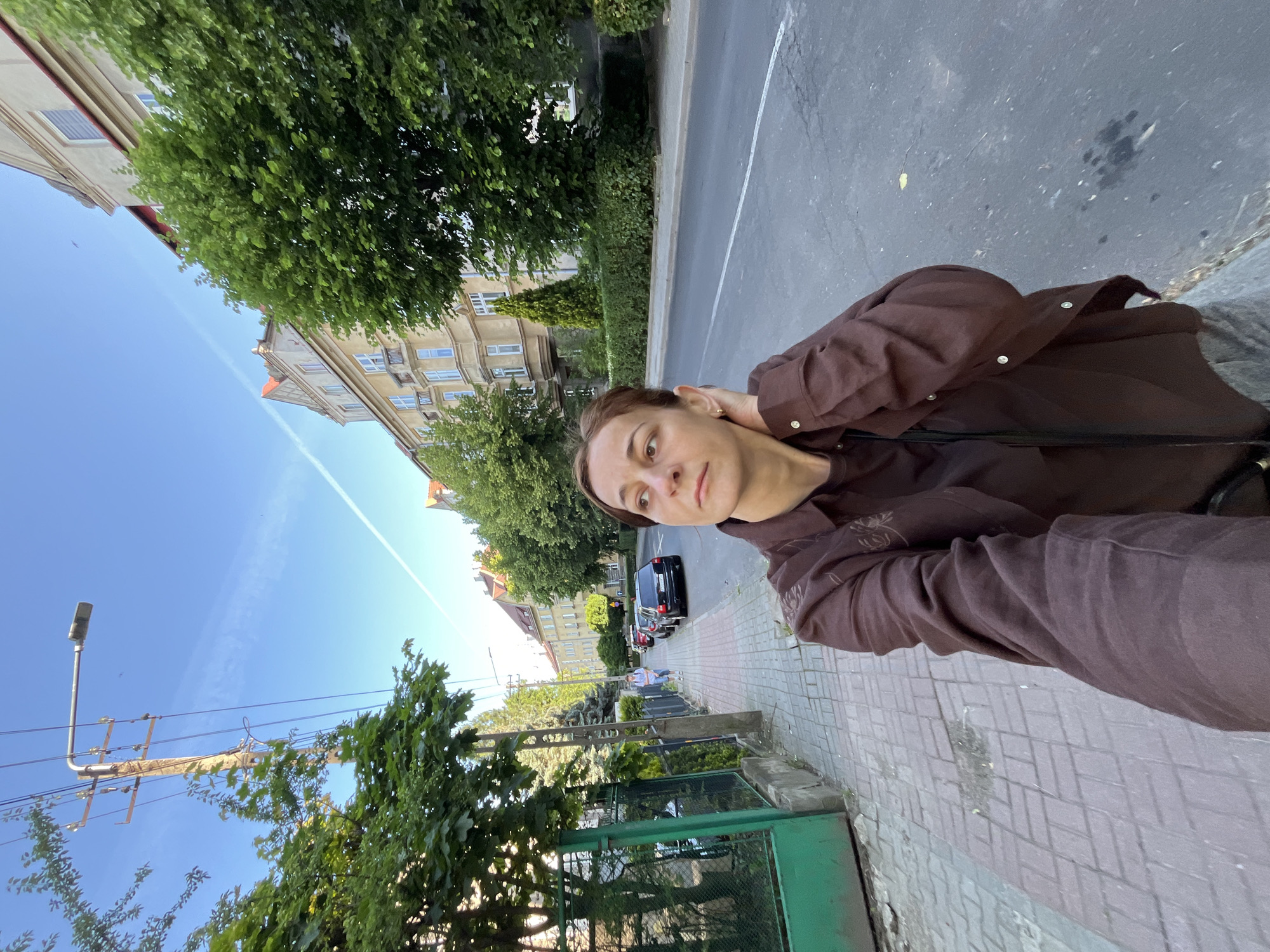 A woman with brown hair is taking a selfie on a street in Unknown, Unknown. She is wearing a brown shirt. She is standing on a sidewalk made of bricks. To her left is a green gate and some trees. To her right is a street with cars parked on the side. In the background, there are buildings and trees. The sky is blue with some white clouds.