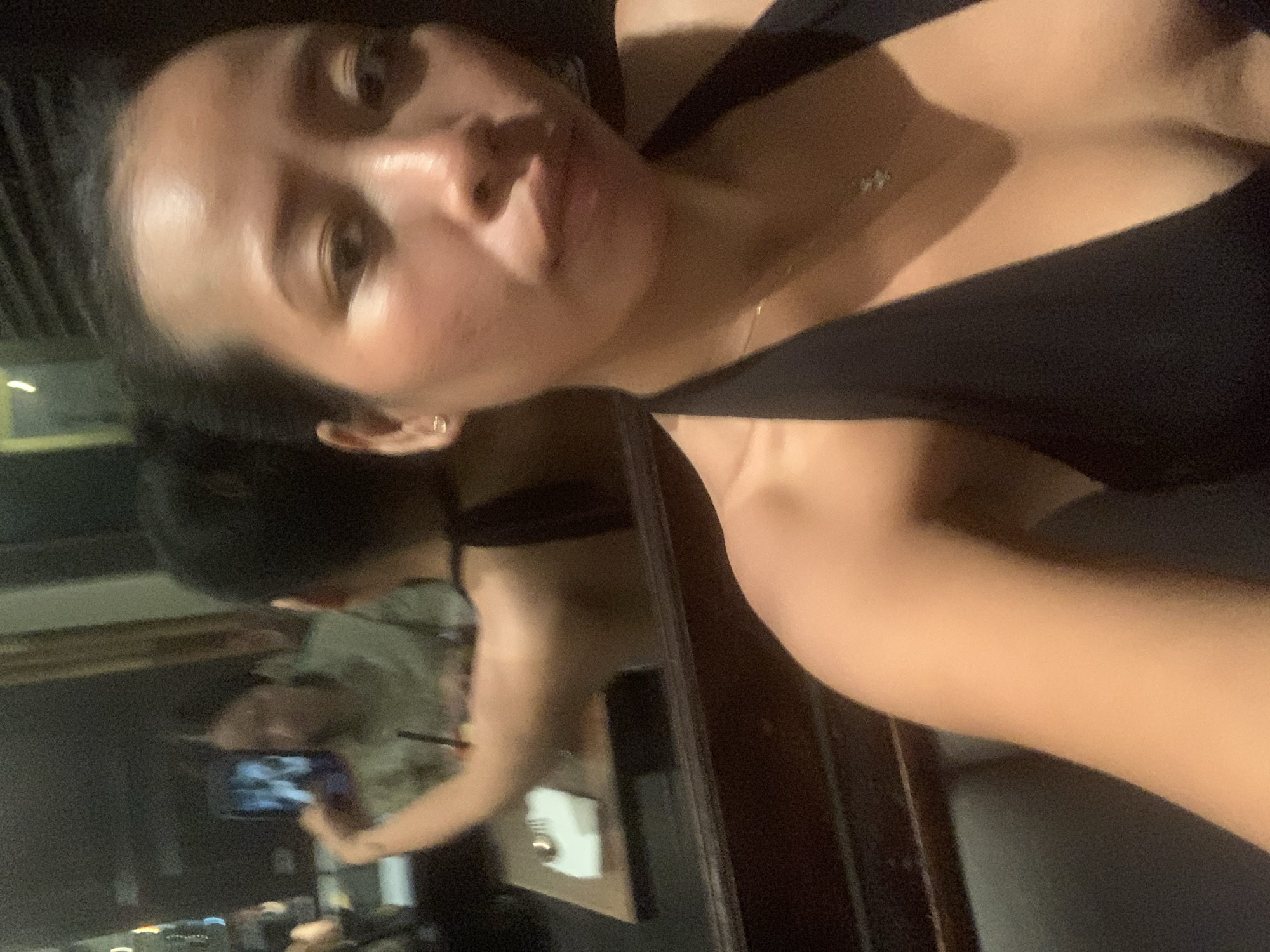 A woman in a black top takes a selfie in a dimly lit indoor location, possibly a restaurant, in North Kuta, Indonesia. Her reflection is visible in a mirror behind her, where another person is holding a smartphone. Other people are also visible in the background of the reflection.
