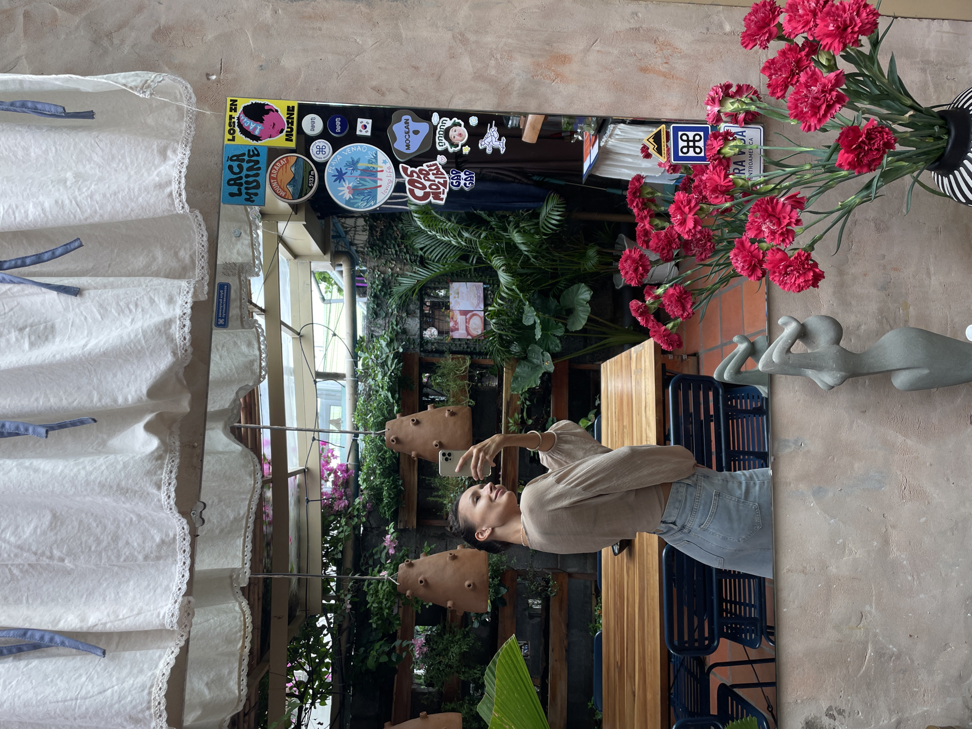 A young woman, seen from the side in a mirror selfie, stands in a cafe or restaurant with lush green plants and a wooden table in the background. She is wearing a beige long-sleeved shirt and light-wash jeans, and her hair is pulled back. She is holding a smartphone in her right hand and smiling. The mirror is decorated with various stickers, including one that reads "LOST IN MUINE" and another with a picture of a person's face. Outside the reflected scene, a bouquet of bright pink carnations in a striped vase is visible in the foreground, alongside a sculpture. The location is identified as Mũi Né, Vietnam.