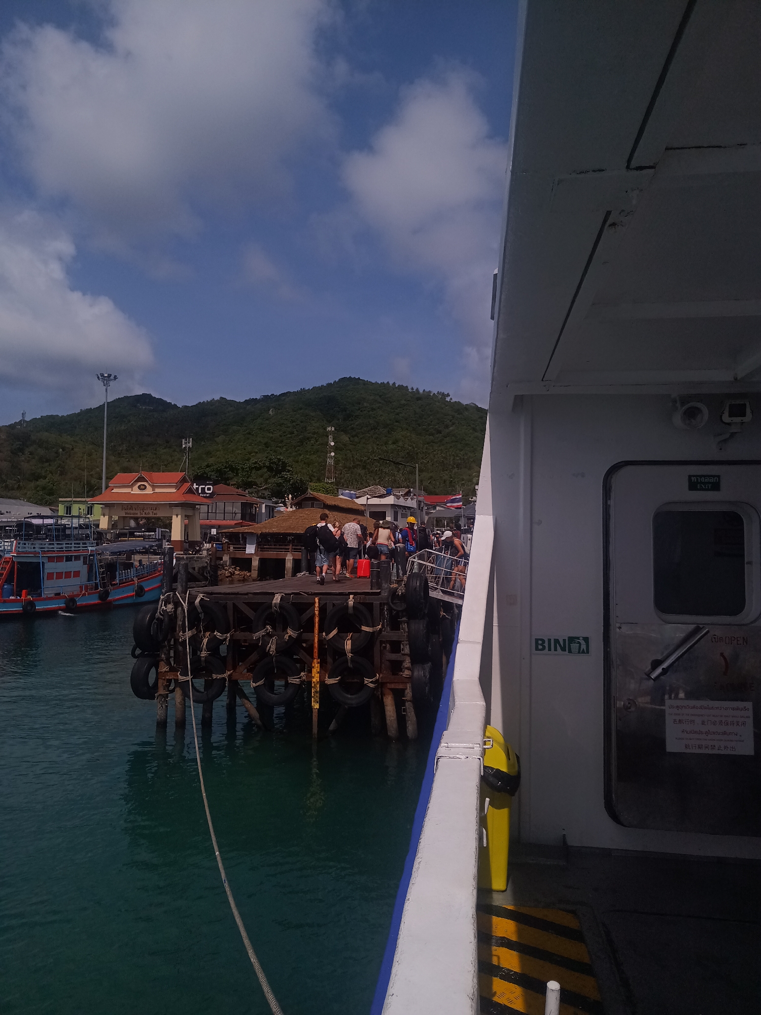The image depicts a ferry docked at a pier in Tambon Ko Tao, Thailand, on a partly cloudy day. The pier is constructed of wood and supported by pilings wrapped with used tires for cushioning. Several people are walking on the pier. Adjacent to the pier, there are traditional Thai-style buildings with red roofs. The foreground of the image is dominated by the side of the ferry, including a white exterior wall with a door, a bin sign, and safety warning in Thai and Chinese languages. The water is a greenish-blue color. In the background, there are lush green hills.