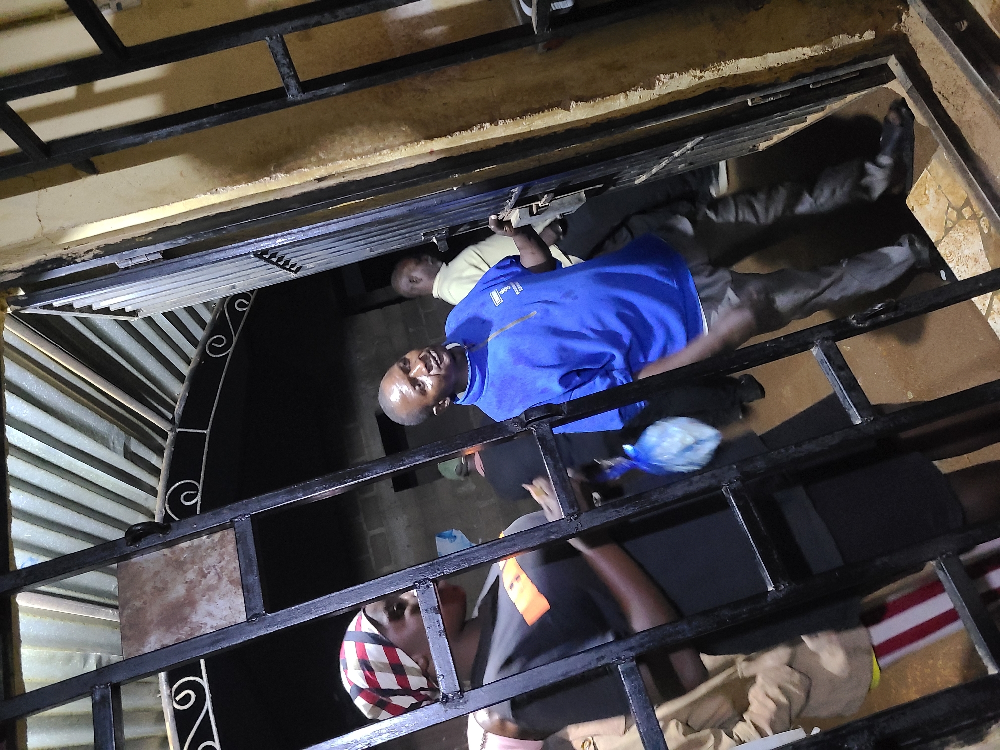 This image captures a multi-level indoor scene, likely a stairwell or an open building structure, viewed from above. The primary subjects are several individuals positioned on different levels, separated by black metal railings that crisscross the frame.
In the foreground and center, a bald man wearing a bright blue short-sleeved shirt looks upwards towards the camera with a neutral expression. He is partially obscured by a horizontal metal bar. Behind him and slightly to his left, another bald man in a light yellow or beige short-sleeved shirt is visible, also looking towards the upper part of the frame, with his hands near another metal bar. Further down and to the right, a person in dark gray pants and dark shoes is seen, possibly descending or standing on a lower step.
Below these individuals, another person wearing a black t-shirt with an orange design and a multi-colored head covering (featuring red, white, and black stripes) is looking upwards, also through the metal railings. Their lower body, dressed in black pants and khaki-colored clothing, is visible at the bottom of the frame. One of the individuals, likely the man in the blue shirt or the person below him, is holding a small, light blue plastic bag.
The setting appears to be a utilitarian building with light-colored, possibly concrete or plastered walls that show some wear and discoloration. The floor visible below the individuals consists of light-colored, possibly tiled, surfaces. The prominent black metal railings are sturdy, some featuring decorative scrollwork and a small, rectangular tan-colored tile embedded within them. The lighting suggests an indoor environment, possibly illuminated by artificial light, given the absence of strong natural light and the general darkness in certain areas. No discernible text is visible on clothing or objects. The individuals appear to be either moving through or gathered within this multi-level structure.