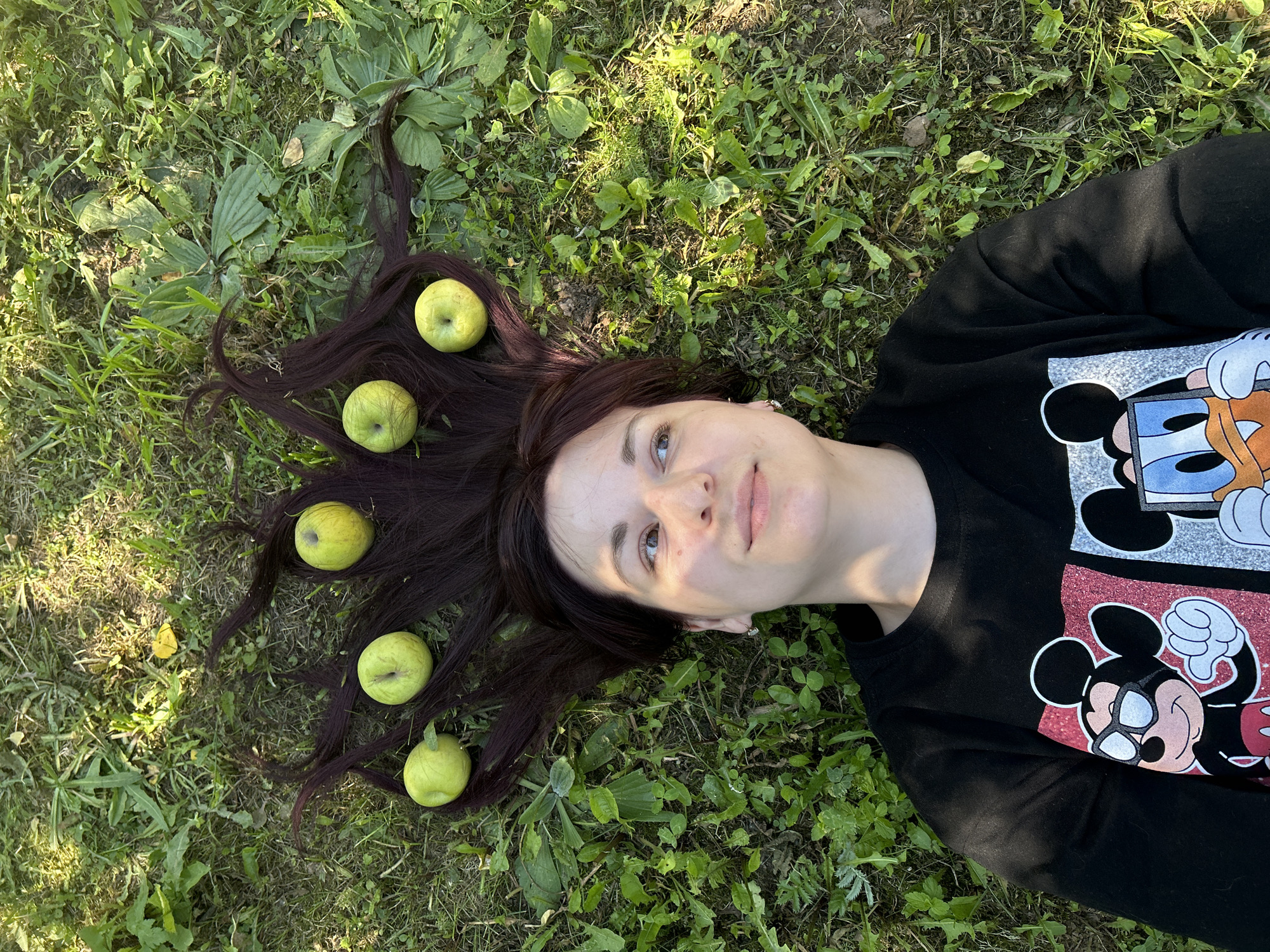 A young woman with dark hair and fair skin is lying on her back in a grassy area, possibly a park or backyard, in the city of Foky, Russia. Her hair is spread out around her head, and several green apples are placed within it. She is wearing a black sweatshirt with images of Mickey Mouse and Donald Duck on the front. The sun is casting shadows on the grass. The scene appears to be set during the day.