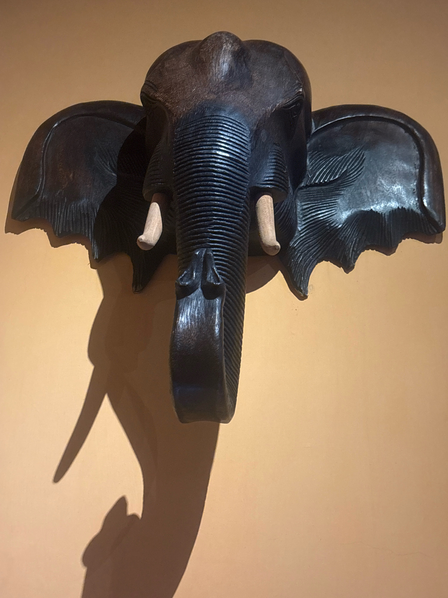 A carved wooden elephant head is mounted on a textured, orange wall. The elephant has large, textured ears, a ridged trunk, and visible tusks. A shadow of the elephant head is cast on the wall to the left, with the shadow of the trunk appearing to curl. The image is captured from a frontal, slightly downward angle. The location of this object in Macau, China, is unknown.