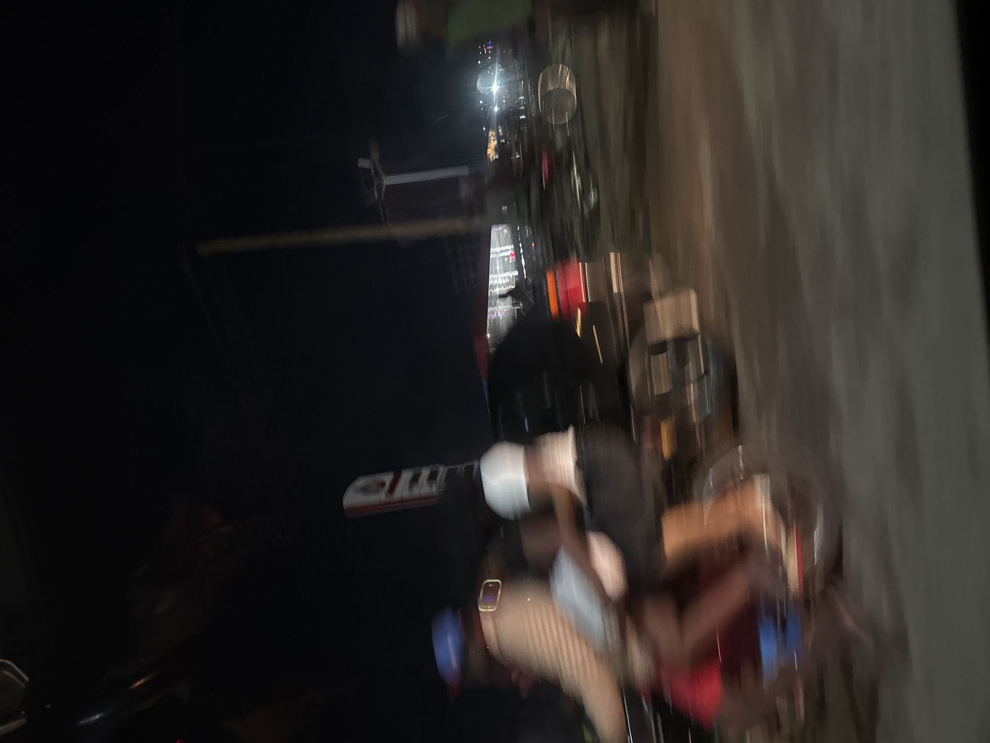 The image, taken at night, is blurred and appears to be from Abuja, Nigeria, judging by the urban infrastructure visible in the background. In the foreground, a person is seen sitting with others, possibly on a mobile food cart or similar structure. Their attire includes a blue head covering and a watch is visible on their arm. The environment is dimly lit with streetlights illuminating the area. The scene is likely a street scene in an urban part of Abuja.