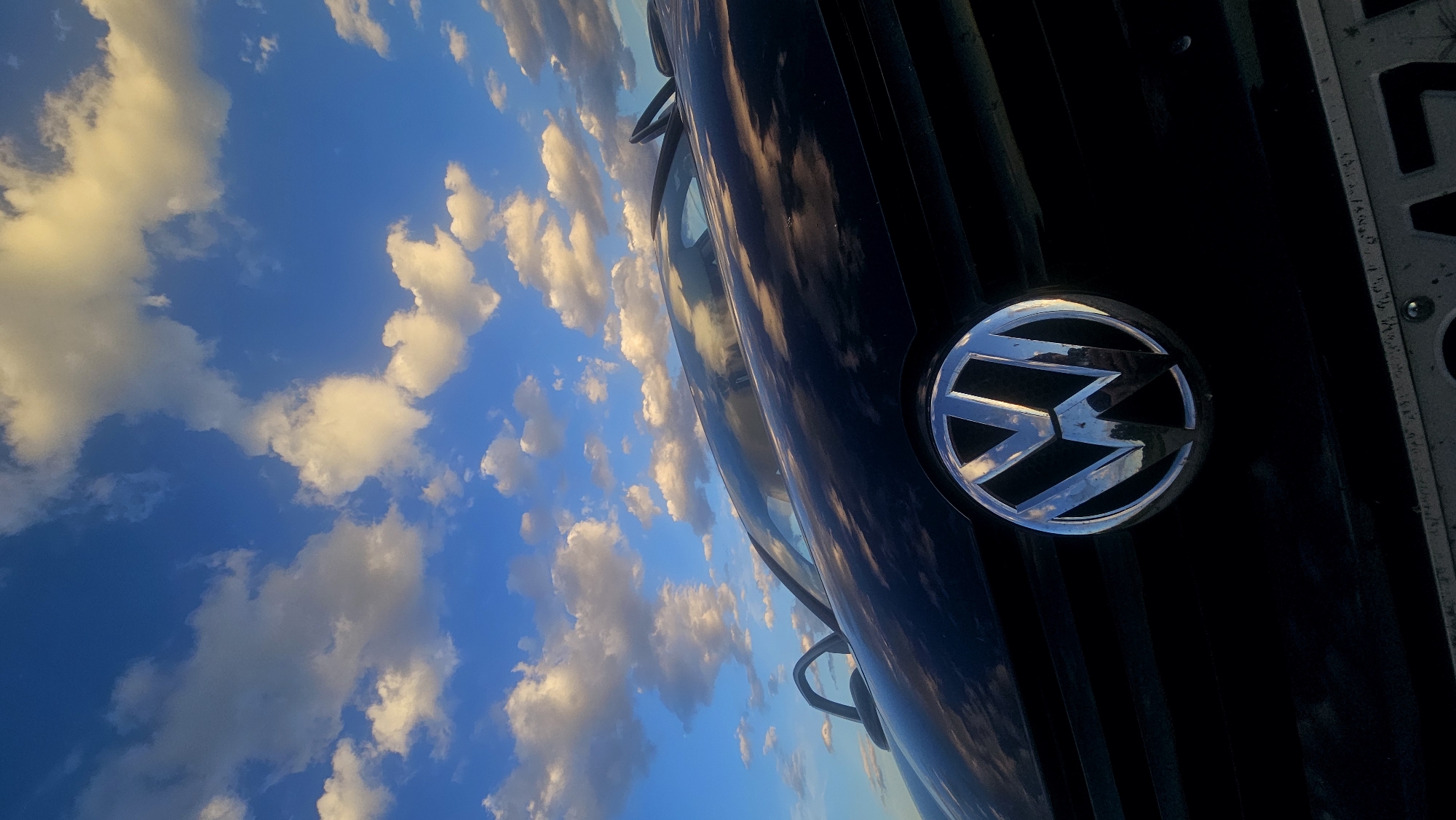 The image shows a low-angle, close-up shot of the front of a black Volkswagen car. The iconic circular Volkswagen logo is prominently displayed in the center of the frame, reflecting the sky. The car's hood and windshield are visible, with the sky and scattered white clouds dominating the upper portion of the image. The scene appears to be outdoors, with the lighting suggesting it might be near sunset or sunrise due to the warm tones in the clouds. There are no people visible in the image, and the environment outside the car is not detailed. The focus is on the car's emblem and the reflected sky, creating a dramatic contrast between the dark vehicle and the bright, cloud-filled sky.