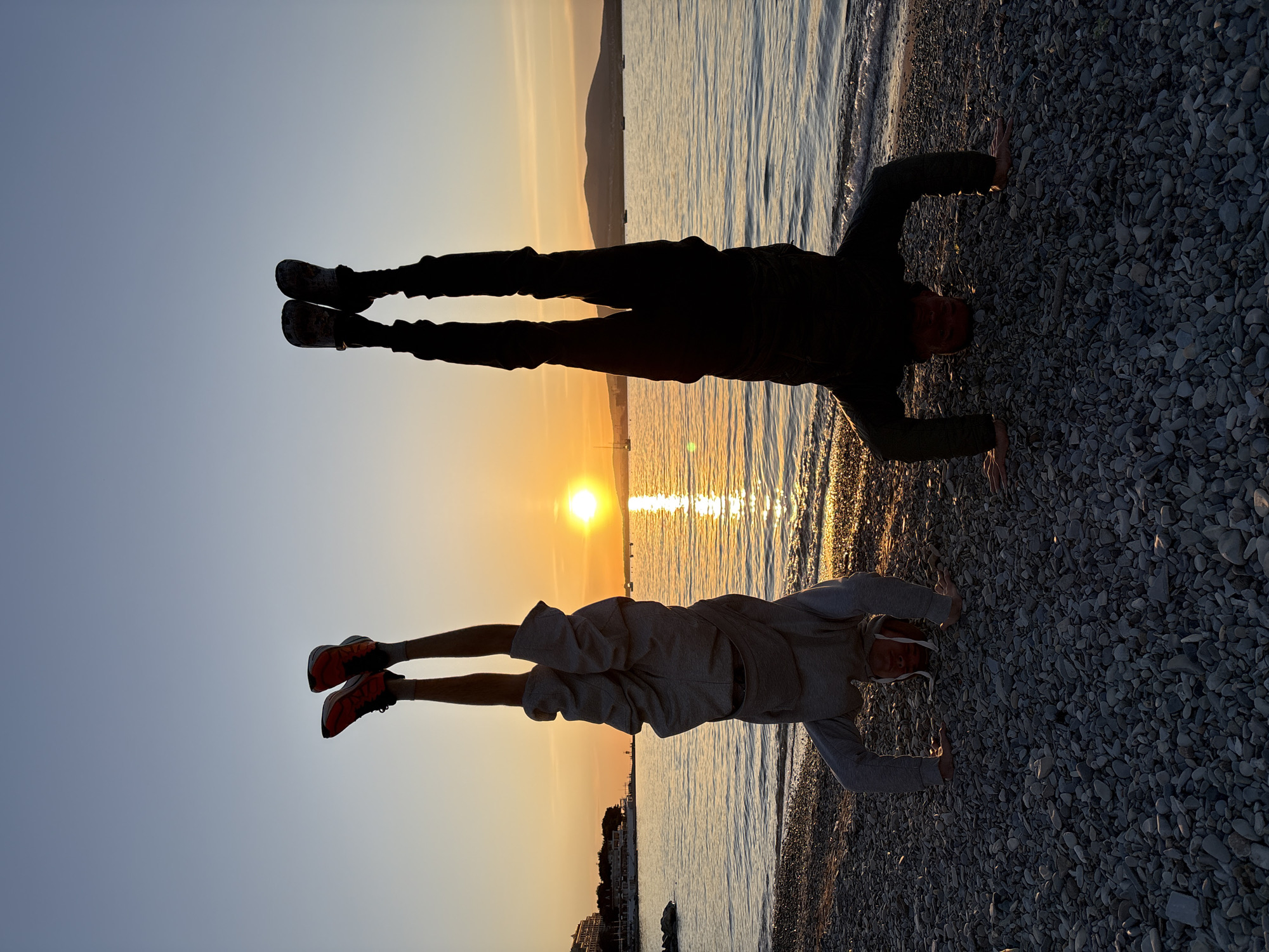 This media file depicts a serene sunset scene on a pebble beach in Gelendzhik, Russia, featuring two individuals performing handstands.
**Subjects:** Two people are the primary subjects, silhouetted against the setting sun. The individual on the left (further from the sun) is wearing dark, long-sleeved clothing and dark shoes. The individual on the right (closer to the sun) is dressed in a light grey hoodie, grey shorts, and distinctive orange/red athletic shoes. The scene also includes the vast expanse of a calm sea or large lake, a pebble beach, and distant mountains or hills on the horizon. A small boat or yacht is visible in the water in the middle ground, and hints of a coastal town or structures can be seen along the distant shoreline.
**Activity or event taking place:** Both subjects are engaged in physical activity, specifically performing handstands on the pebbled shore. This suggests exercise, acrobatics, or a playful moment captured during a scenic sunset.
**Notable details:** The time of day is clearly sunset, with the sun appearing as a bright, golden orb low on the horizon, casting a brilliant reflection across the water. This warm, golden light backlights the subjects, creating strong, dramatic silhouettes. The sky transitions from a soft orange near the horizon to a clear, pale blue higher up. The water is calm with gentle ripples near the shore. The pebble beach is distinctively textured. No visible text is present in the image. The location is confirmed as Gelendzhik, Russia, aligning with the coastal features of the image.