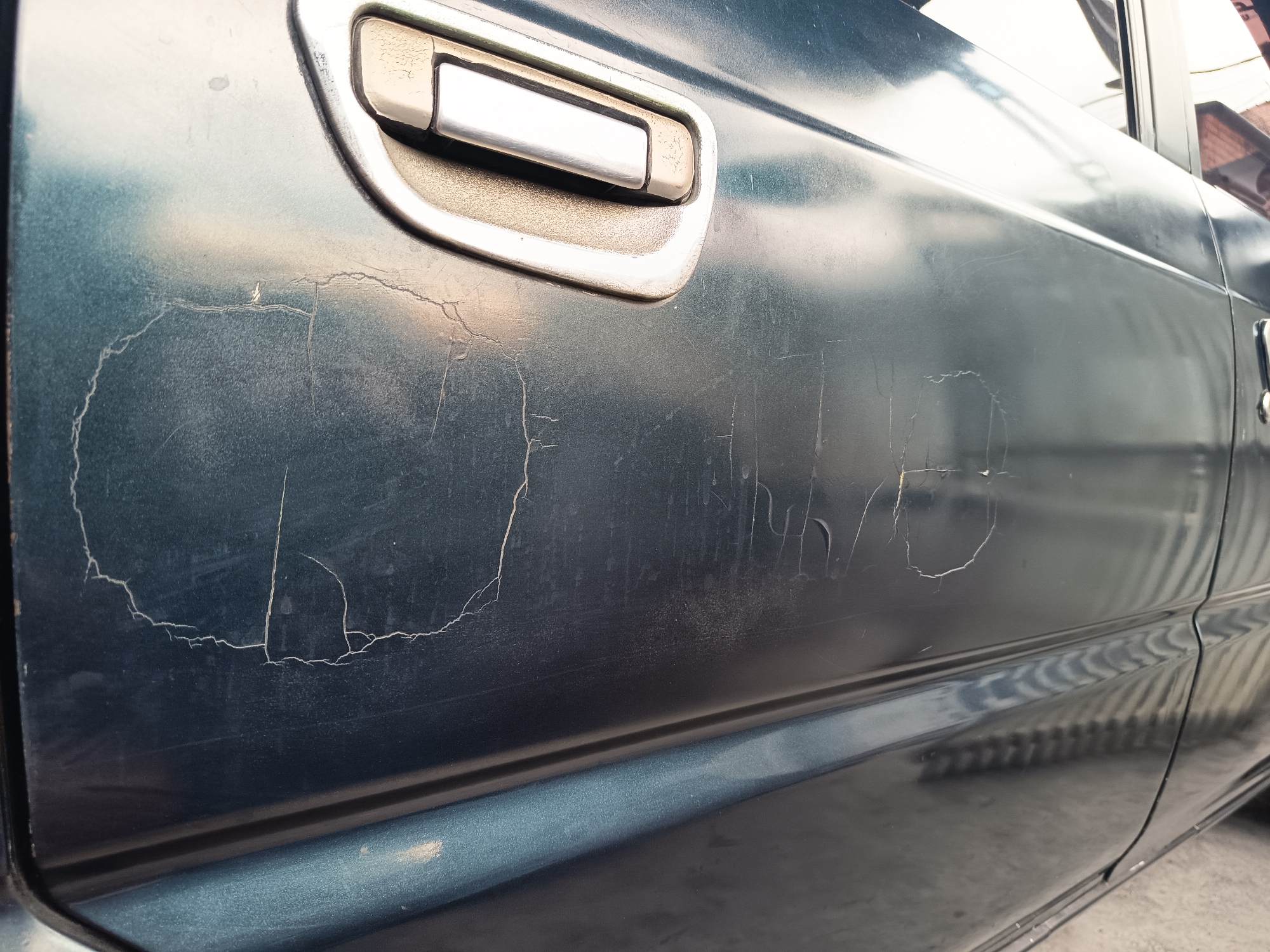 This image shows a close-up view of the side of a dark teal-colored vehicle, specifically the driver's side door. The paint on the car is significantly deteriorated, exhibiting large areas of cracking and peeling, particularly on the lower half of the door. Several scratches are also visible across the surface. A chrome-accented door handle is present on the upper portion of the door. The surrounding environment is not clearly visible, but a light-colored surface, possibly concrete, is visible at the bottom of the frame, suggesting an outdoor setting. There are no people or animals present in the image. The image appears to have been taken in an urban or suburban environment, likely a parking area or street. The specific location within Sugio, Indonesia, cannot be determined from the visual information alone.