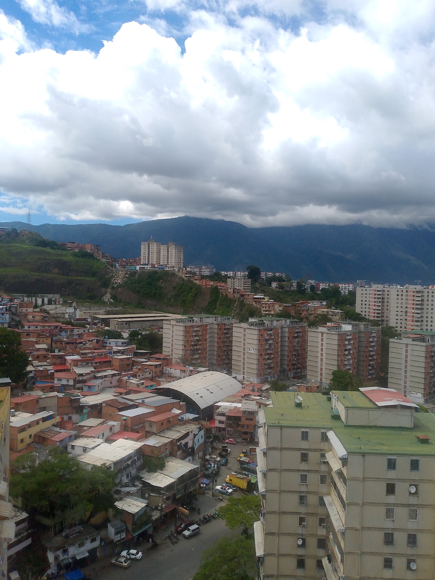 The image captures a dense urban landscape in Caracas, Venezuela, characterized by a mix of residential buildings and a mountainous backdrop. In the foreground, a cluster of multi-story apartment buildings, primarily with beige or light-colored facades and numerous windows, dominate the view. Some buildings feature visible balconies and satellite dishes. The roofs of many buildings are flat, some covered in green or red material. Interspersed with these larger buildings are numerous smaller, densely packed houses with red tile roofs, indicative of a shantytown or informal settlement, built on the steep hillside. The contrast between the modern apartment blocks and the more rudimentary housing is striking.
A road network is visible winding through the lower-lying areas, with several vehicles, including cars and motorcycles, parked or in motion. The environment is partly cloudy, with large, white cumulus clouds against a blue sky, and darker, more ominous clouds gathering over the mountains in the distance. The mountains, covered in vegetation, form a dramatic natural boundary to the city. A large, white, arched structure, possibly a covered market or sports facility, is also present within the urban sprawl. The scene suggests a vibrant, if somewhat chaotic, urban environment with a clear socio-economic stratification visible in the housing types.