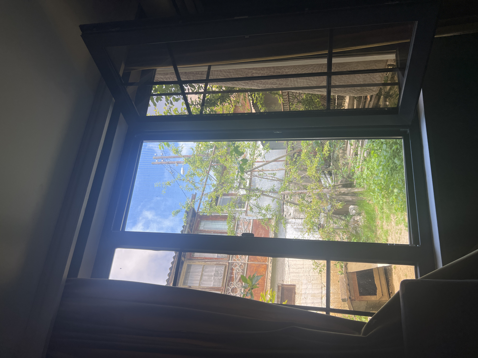 The image presents a view from inside a dark room looking out through an open window, in the city of Unknown, Unknown. Dark beige curtains frame the left side of the window, partially obscuring the view. The window itself has dark-framed panes, with the rightmost vertical pane open inwards. Beyond the window, a bright outdoor scene is visible under a blue sky with scattered white clouds, contrasting with an overcast, lighter sky section on the upper left. Directly outside, a multi-story structure is present, featuring a lower level constructed of light-colored, possibly stuccoed material, and an upper level with a reddish-brown facade and an intricate white metal railing or decorative element. Several windows with white frames and white lace curtains are visible on the upper level. A prominent green tree with sparse foliage stands in the center, and various other green plants and grasses populate the ground level. To the far left, a makeshift doghouse or shelter built from concrete blocks and wood/metal is situated on sandy ground. Utility poles and power lines are visible against the sky and interspersed with the vegetation. The scene depicts a densely built, somewhat overgrown backyard or alleyway environment.