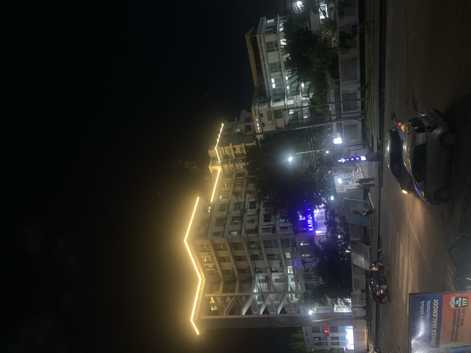 The image depicts a night scene in Dar es Salaam, Tanzania. Prominently featured is a tall, modern building adorned with yellow, zig-zag shaped decorative lighting. The building is labeled "KAIRU" in blue lettering. The street in front of the building is illuminated by streetlights. A silver car is parked on the side of the road. Other vehicles and possibly a motorbike can be seen. A sign promoting HIV testing and prevention with the Swahili phrase "TIMIZA MALENGO!" and a toll-free number is visible in the foreground. The image is taken at night, and the surrounding sky is dark.