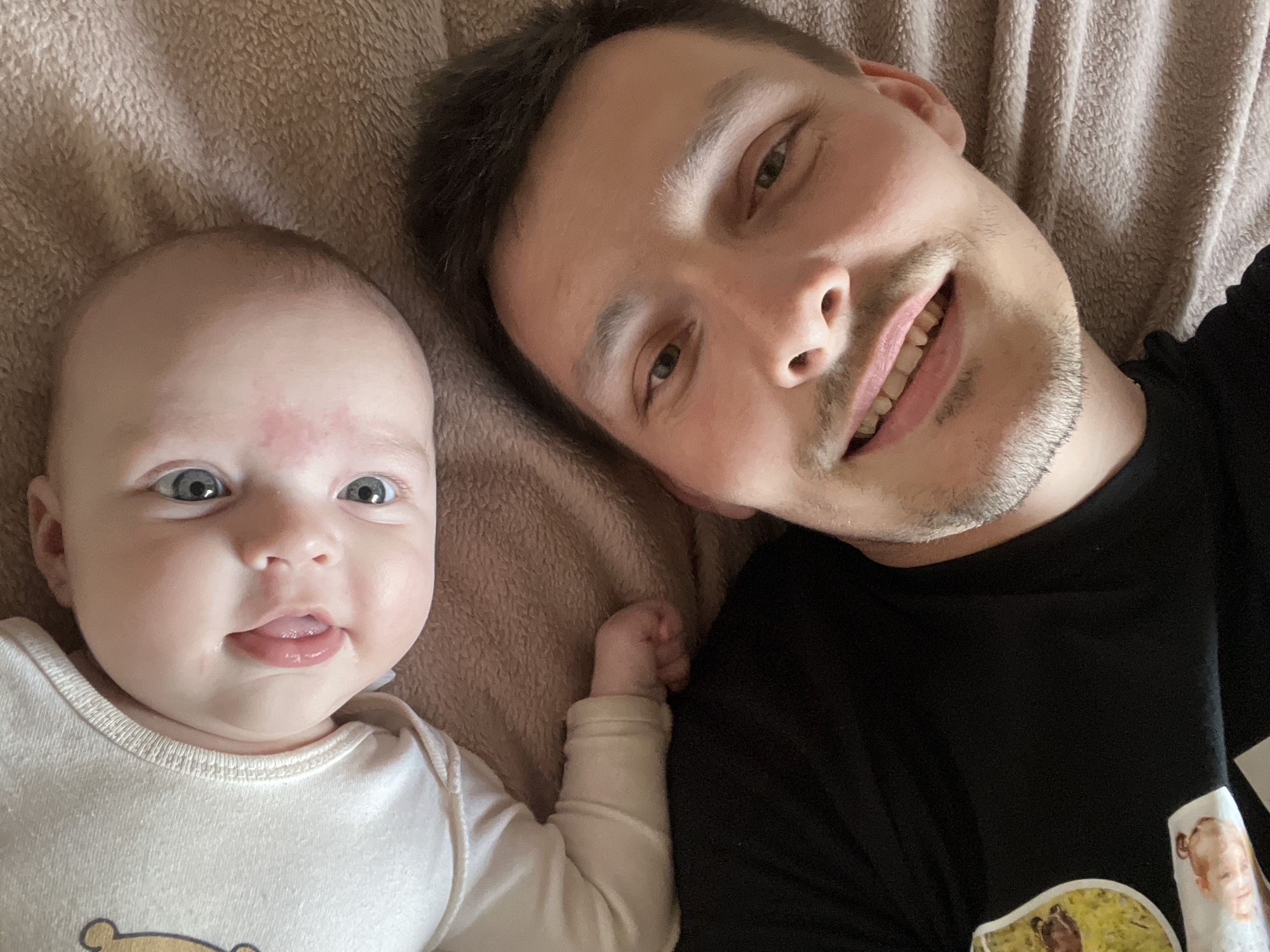 A selfie depicts a fair-skinned baby and a man lying on what appears to be a tan-colored blanket or bedspread. The baby, wearing a white long-sleeved onesie with a cartoon bear design, is on the left side of the frame, with its tongue slightly protruding. The man is on the right side of the frame, wearing a black t-shirt. The man has short brown hair, light stubble, and is smiling, looking towards the camera. The background is out of focus, but the blanket or bedspread has a soft, textured appearance. There is no information to suggest the location is in the city of Chaikovsky, Russia.