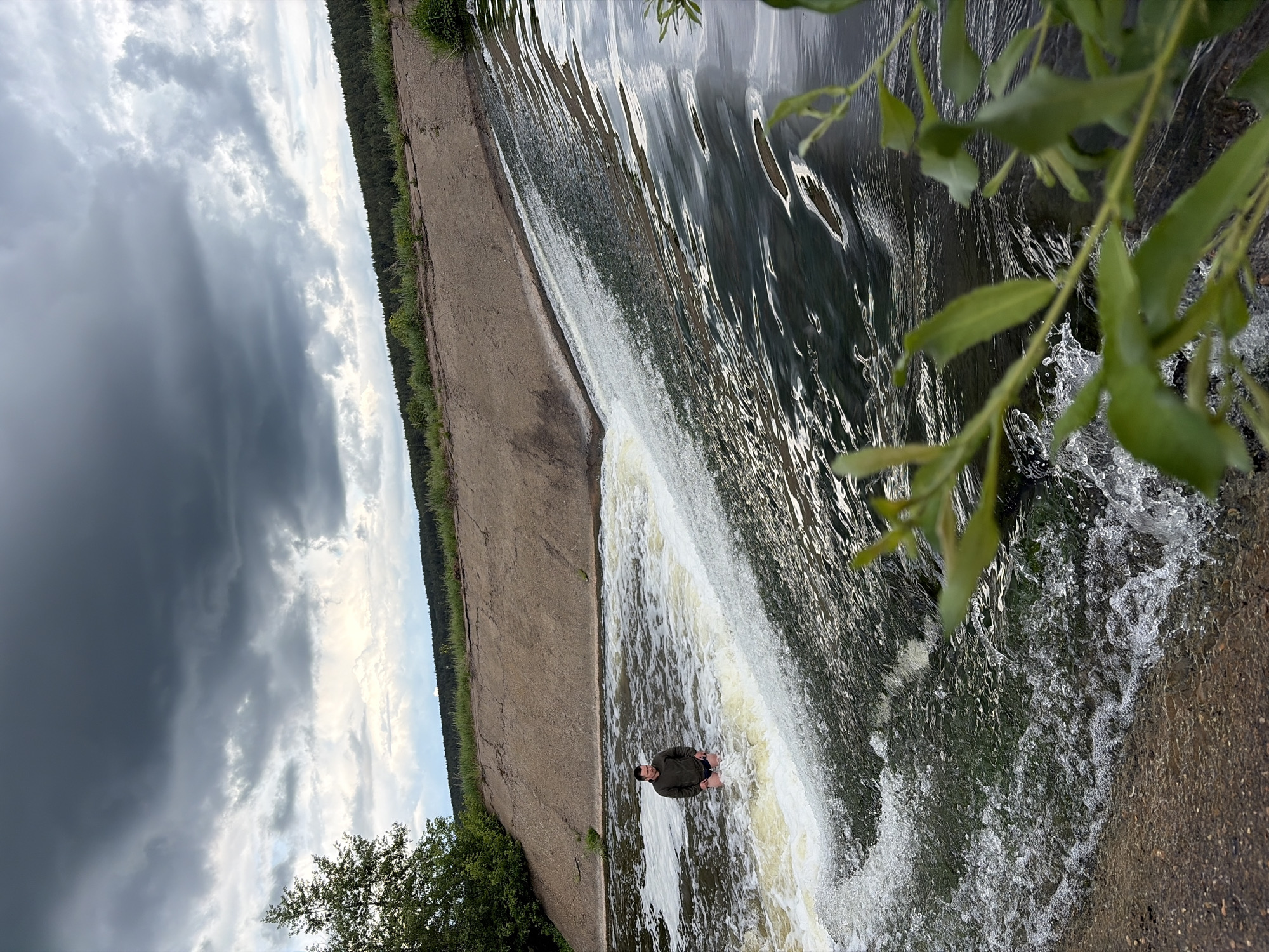 A man stands at the base of a small artificial waterfall in what appears to be Nizhnii Vishur, Russia. The waterfall is created by a dam or weir structure, and the man is standing in the cascading water. The background shows a concrete structure with vegetation along the edge, along with a treeline and a cloudy sky. A leafy branch is in the extreme foreground, partially obscuring the upper right.