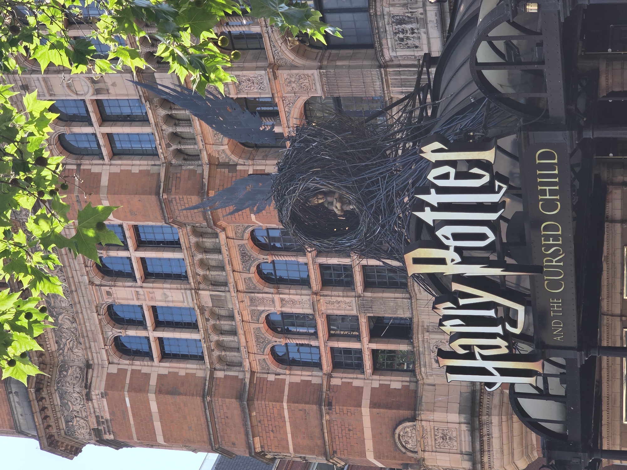The image shows the exterior of the Palace Theatre in London, United Kingdom, displaying the sign for "Harry Potter and the Cursed Child". The sign consists of a large bird's nest sculpture with wings, containing figures in the center. The text "Harry Potter" is displayed vertically in white and gold lettering, while "AND THE CURSED CHILD" is displayed horizontally in gold lettering on a black background. The building itself has a brick facade with ornate architectural details and arched windows. Green leaves from a tree are visible in the foreground.
