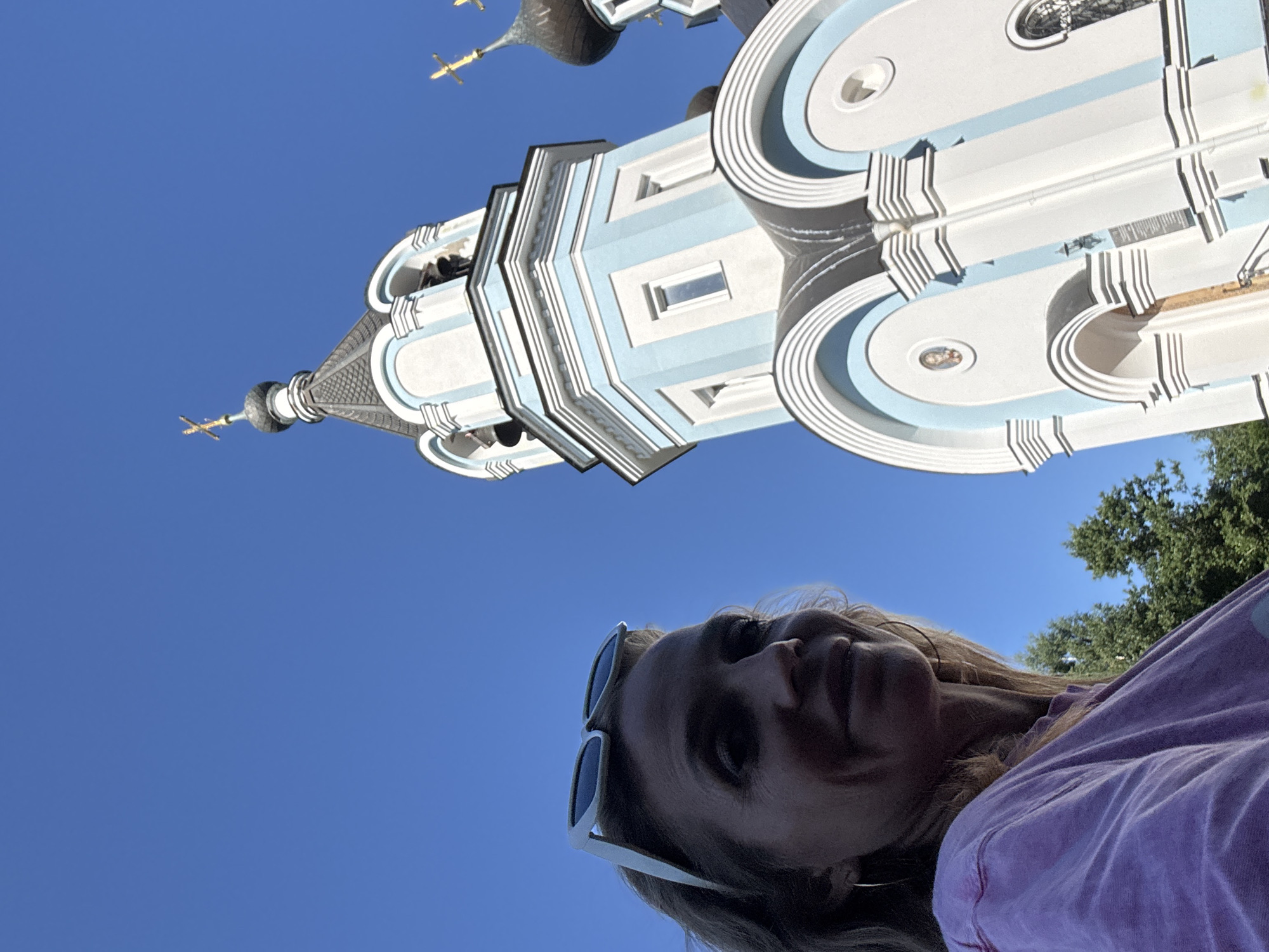 In St. Ozerki, Russia, on a clear day with a bright blue sky, a woman with blonde hair is captured in a low-angle selfie. She is wearing white sunglasses perched on her head and a light purple t-shirt, with a hoop earring visible in her left ear. The background features a white and light blue church with onion domes and golden crosses. The church's architecture includes arched windows and decorative detailing. Trees are visible behind the woman and to the left of the church.