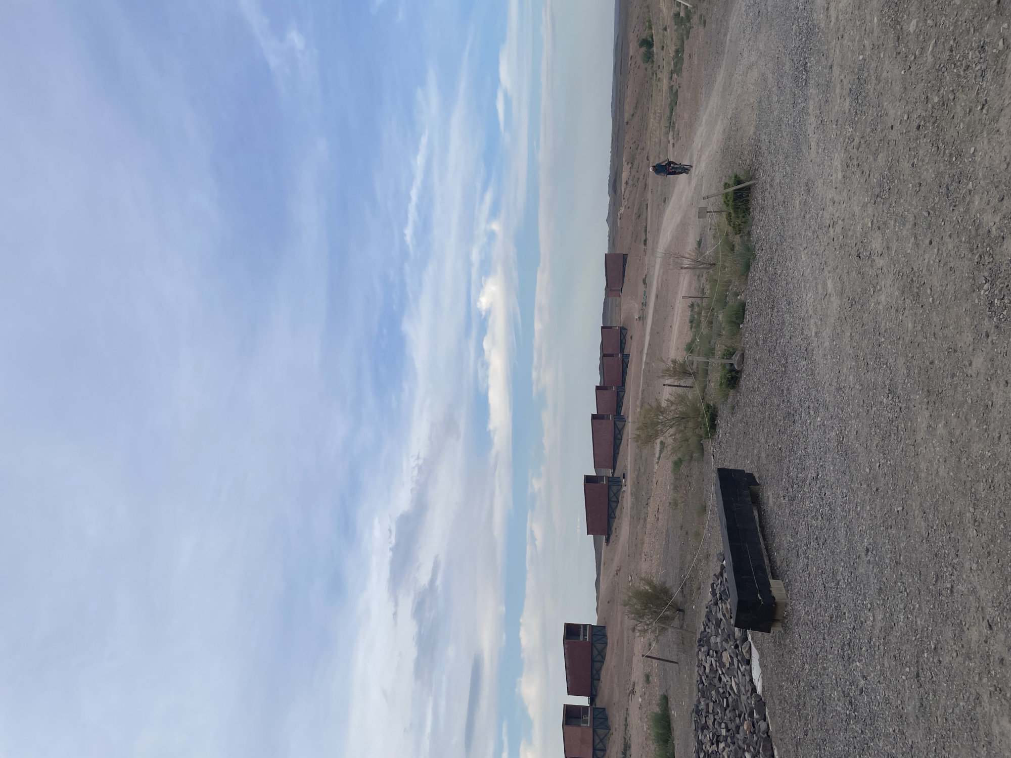 In Tamerlik, Kazakhstan, a person is riding a bicycle on a dirt road. The road curves to the right, leading into the distance. To the left of the road, several red rectangular structures are visible, set on stilts in a barren, arid landscape. These structures appear to be dwellings or cabins. In the foreground, a black bench made of wood sits on a gravelly surface, next to a pile of stones. The sky above is a pale blue with scattered white clouds. The overall environment is dry and sparsely vegetated, with low bushes and dry grasses.