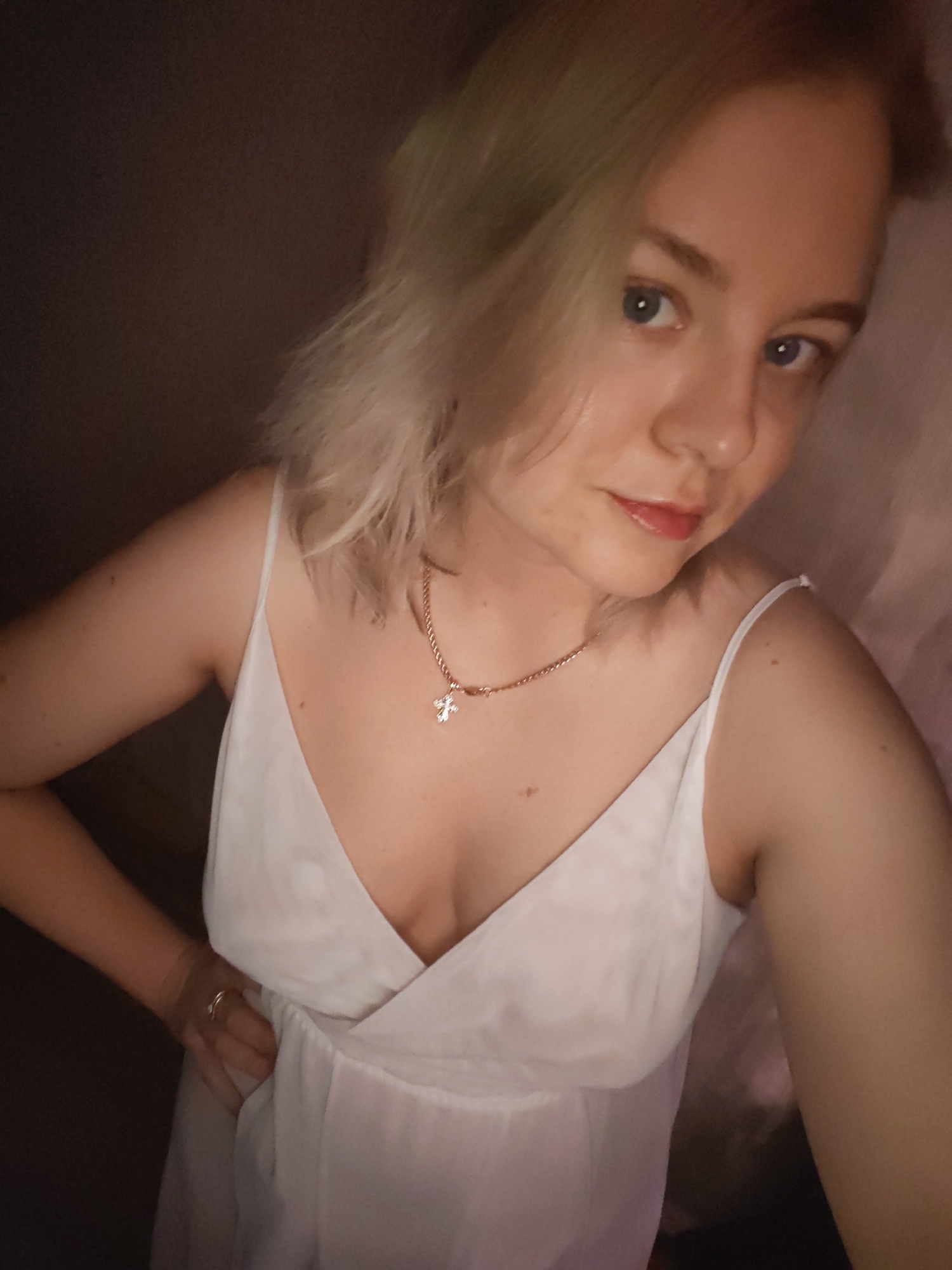 A woman with blonde hair and blue eyes is posing for a selfie. She is wearing a white dress with thin straps and a gold necklace with a cross pendant. Her left hand rests on her hip, and she is wearing a ring on her left ring finger. The background is dark and indistinct. The location is unknown and cannot be determined from the image.