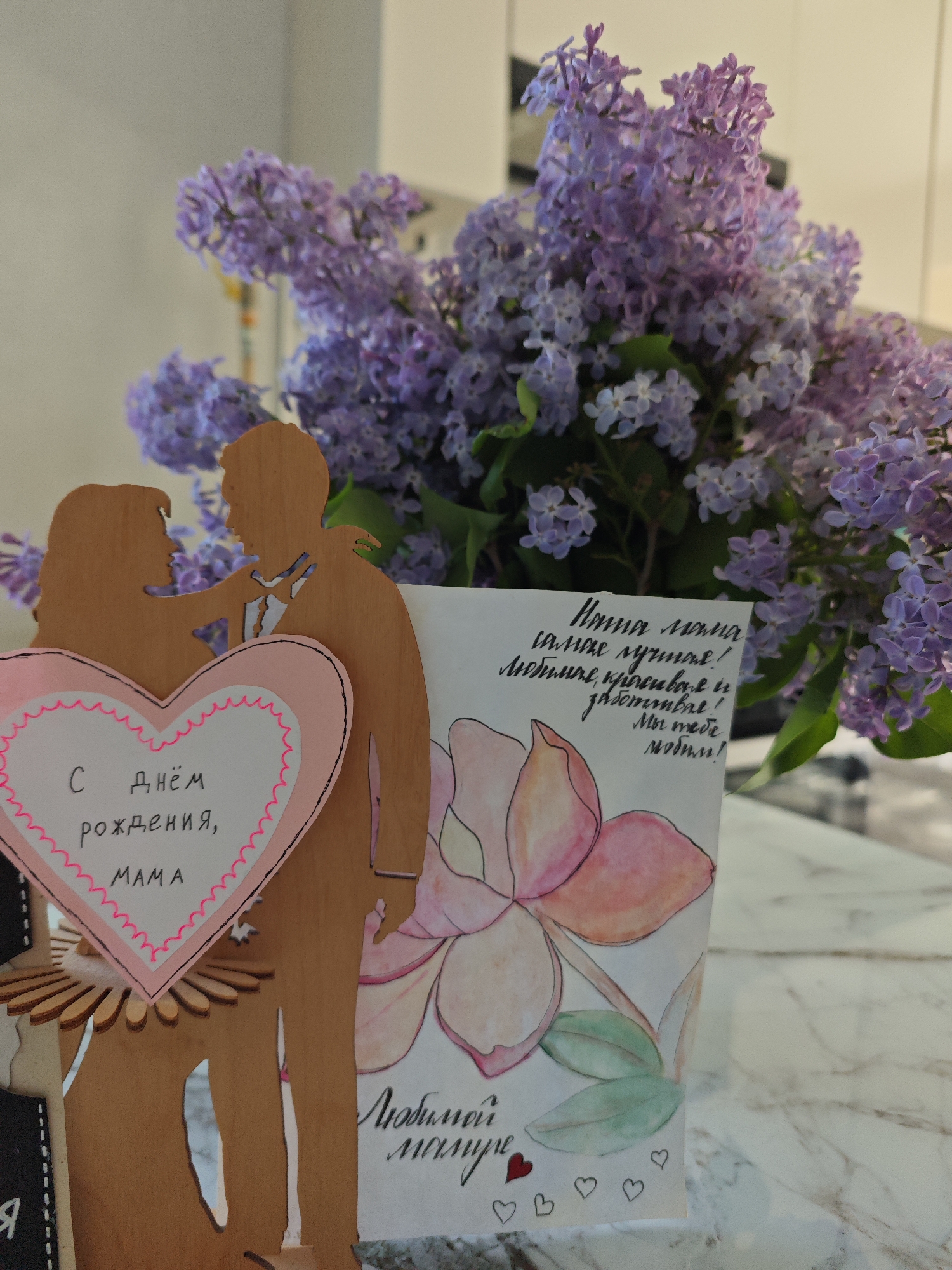 A still life shot features a bouquet of purple lilac blooms. Placed in front of the flowers are two cards or gifts. One is a wooden cutout of a couple, with a heart attached displaying the Russian text "С днем рождения, мама" (Happy Birthday, Mom). The other card is a watercolor-style drawing of a flower, with handwritten Russian text that includes "Наша мама самая лучшая!" (Our mom is the best!) and "Любимой мамуле" (To our beloved mom). The background is a blur of what seems like a kitchen or dining area with a marble-like surface. The location, Фоки, Russia, is not discernible from the image's content.