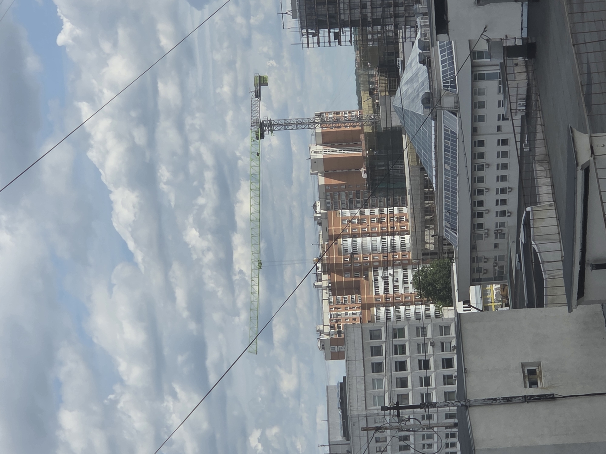 This is a distant, eye-level, outdoor, daytime shot of a cityscape in Moscow, Russia, featuring a construction site dominated by a large tower crane. The crane, a pale green color, extends horizontally across the upper middle portion of the frame, its boom reaching towards the right. It is supported by a tall, lattice-like metal tower.
Beneath the crane and in the background, a cluster of multi-story buildings is visible. The most prominent of these is a tall, orange brick residential building with numerous windows arranged in a grid pattern. To its left, a lighter colored, modern building with a repetitive window design stands. Further to the right, the skeletal structure of another building under construction, clad in scaffolding, is visible.
In the foreground, several more buildings are present. The closest structures appear to be lower-rise, flat-roofed buildings. One building, a light grey color, dominates the bottom left of the frame, featuring a simple facade with a few small, dark windows. Another building to its right has a distinctive, large, multi-faceted glass roof that resembles a pyramid. Power lines and utility poles crisscross the scene, adding to the urban feel. The sky is partly cloudy with a mixture of white and grey clouds against a pale blue background. No people are clearly visible in the frame.