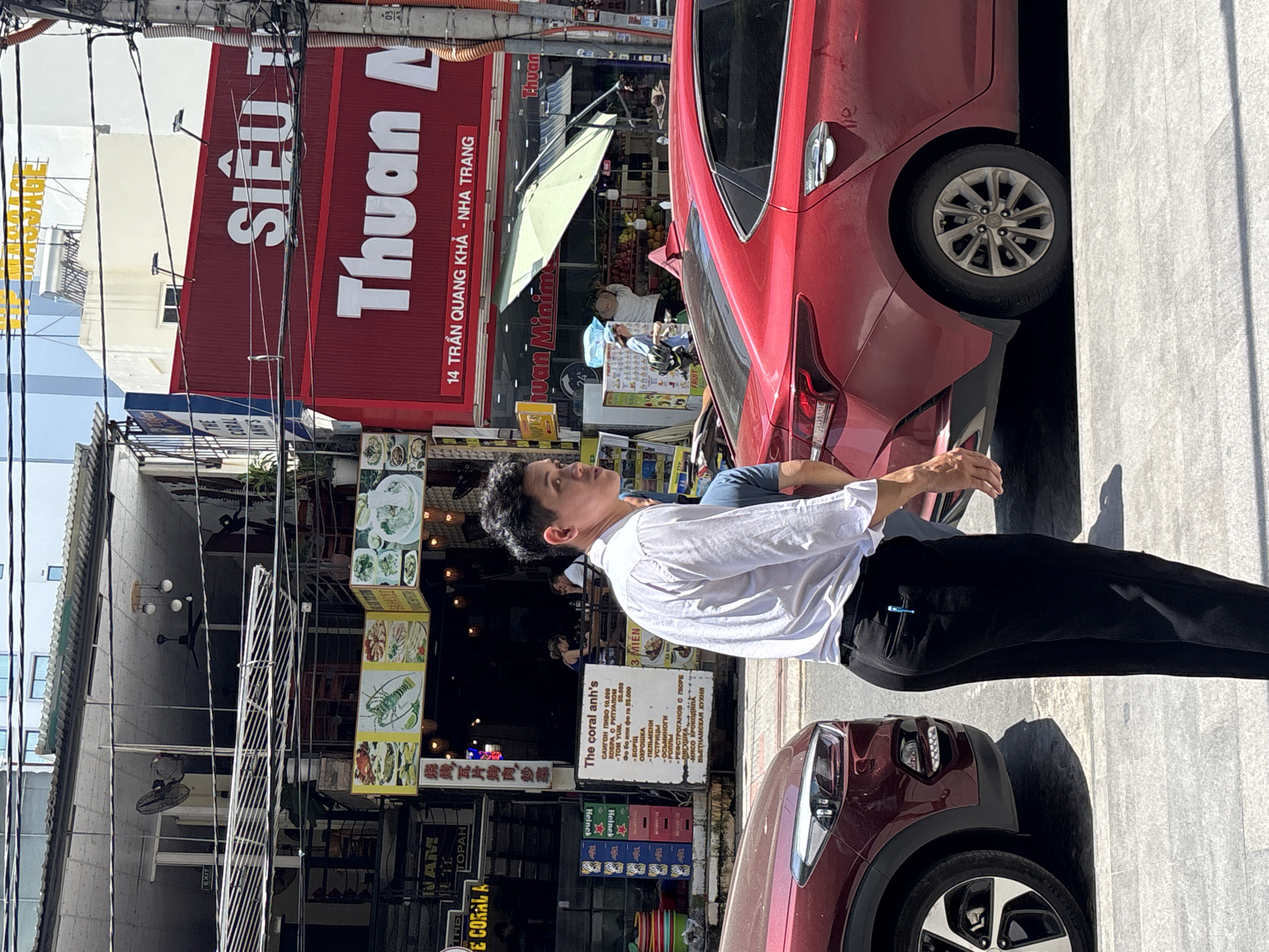 The image depicts a street scene in Nha Trang Ward, Vietnam. A man is standing on the sidewalk next to two parked cars, a red hatchback and a burgundy SUV. Behind him is a row of buildings with signs in Vietnamese and English. One sign reads "SIÊU T Thuan A" and another reads "The coral anh's". The street is lined with power lines. People can be seen walking and sitting in the background.
