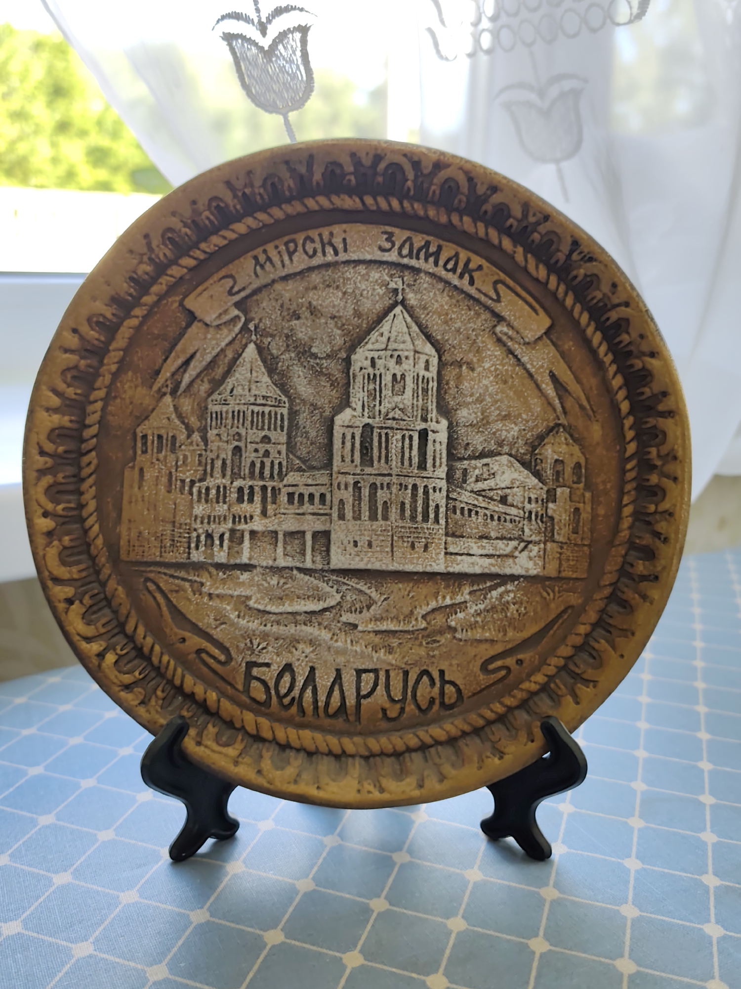 A brown, circular decorative plate rests on a black stand. The plate features a raised relief of the Mir Castle Complex, a UNESCO World Heritage site in Belarus. The text "МІРСКІ ЗАМАК" (Mir Castle) is inscribed in an arc above the castle depiction, and "БЕЛАРУСЬ" (Belarus) is written below. The background of the plate is textured to resemble stone. The plate is positioned on a light blue surface with a white checkered pattern. Behind the plate, a sheer white curtain with a subtle embroidered floral pattern is visible, with green foliage and bright sunlight filtering through from an unseen window.