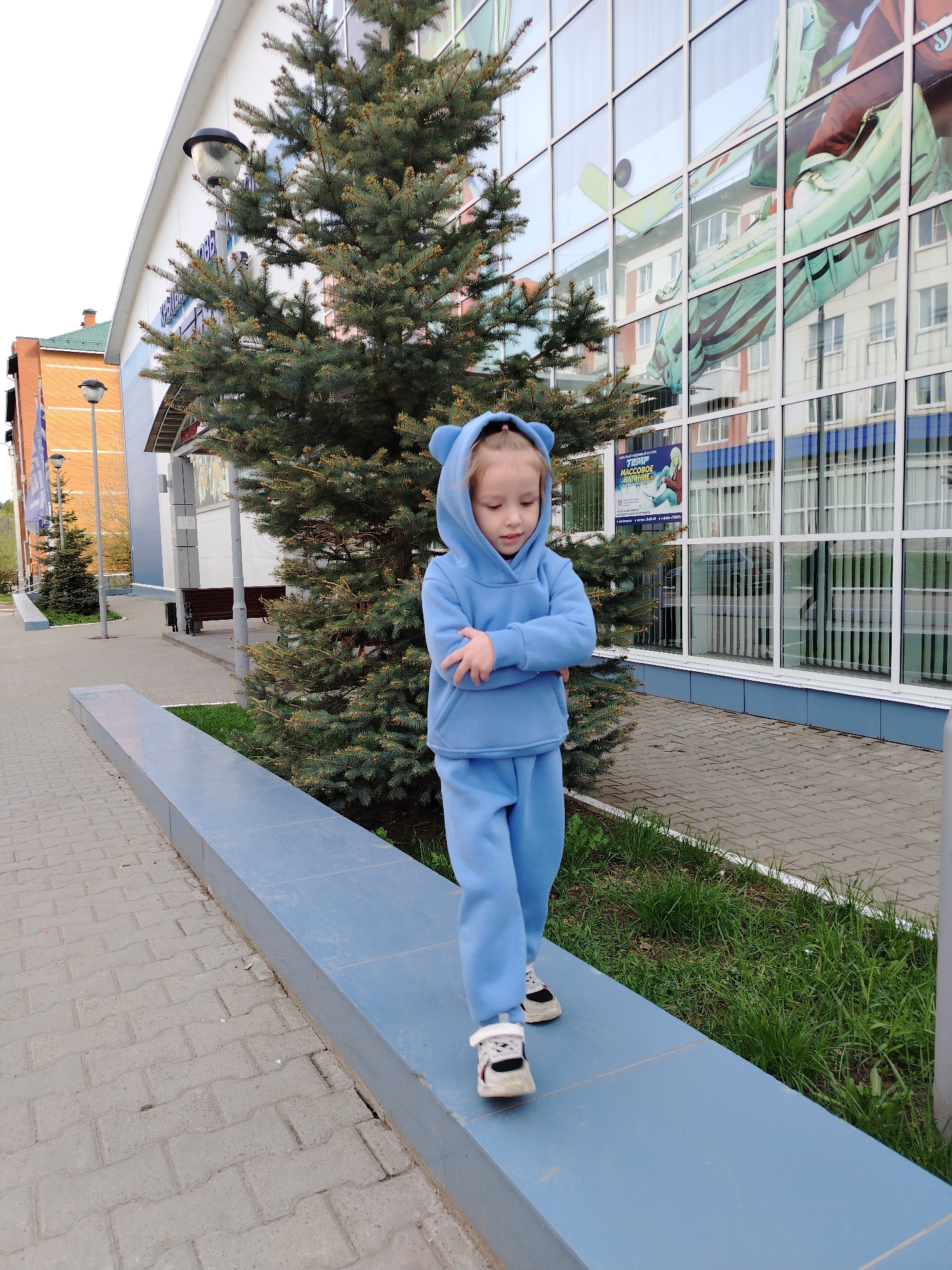 A young girl, dressed in a light blue tracksuit with bear ears on the hood, stands on a raised blue platform, with her arms crossed. She is looking downwards and walking. The girl is wearing sneakers. Behind her is a small evergreen tree. To the right is a building with large, reflective windows that are reflecting other buildings. To the left is a sidewalk made of gray rectangular pavers and a brick building in the background. The location is Chaikovsky, Russia.