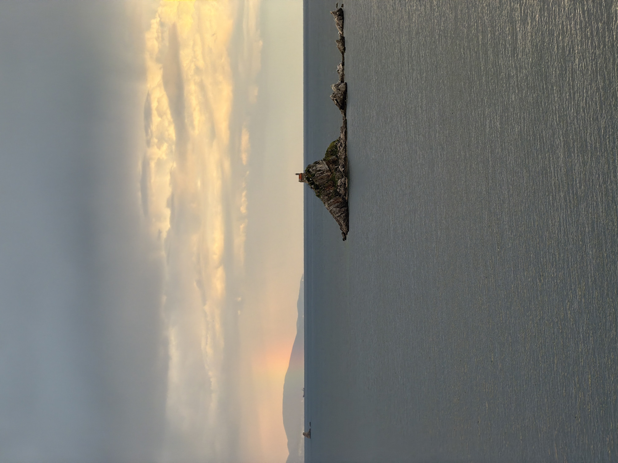 An outdoor landscape photograph captures a serene yet dramatic coastal scene, likely during sunrise or sunset, in Mirište, Montenegro. The image is composed with a clear horizon line, dividing a textured body of water in the foreground from a dynamic sky above.
The sky, occupying the upper half of the frame, is filled with a mix of dark, grey clouds and brightly illuminated, golden-orange clouds, indicating either the early morning or late evening light. Below the sky, the calm water appears dark grey-blue with subtle ripples and reflections, suggesting a tranquil surface.
Along the horizon, a series of rocky islets or a rugged promontory stretches from the left towards the center. The most prominent part of this landmass features some green vegetation and a small, reddish-brown structure, possibly a lighthouse or a remote building. In the far distance, a faint, colorful arc resembling a rainbow is visible, casting subtle hues over a hazy mountain range or distant hills. A few indistinct, tiny shapes, possibly boats, can be discerned on the water near the distant landmass.
No specific human activity or identifiable people are depicted. There is no visible text. The time of day is clearly sunrise or sunset due to the warm, golden light, and the presence of the faint rainbow suggests atmospheric moisture. The scene evokes a tranquil and contemplative mood.