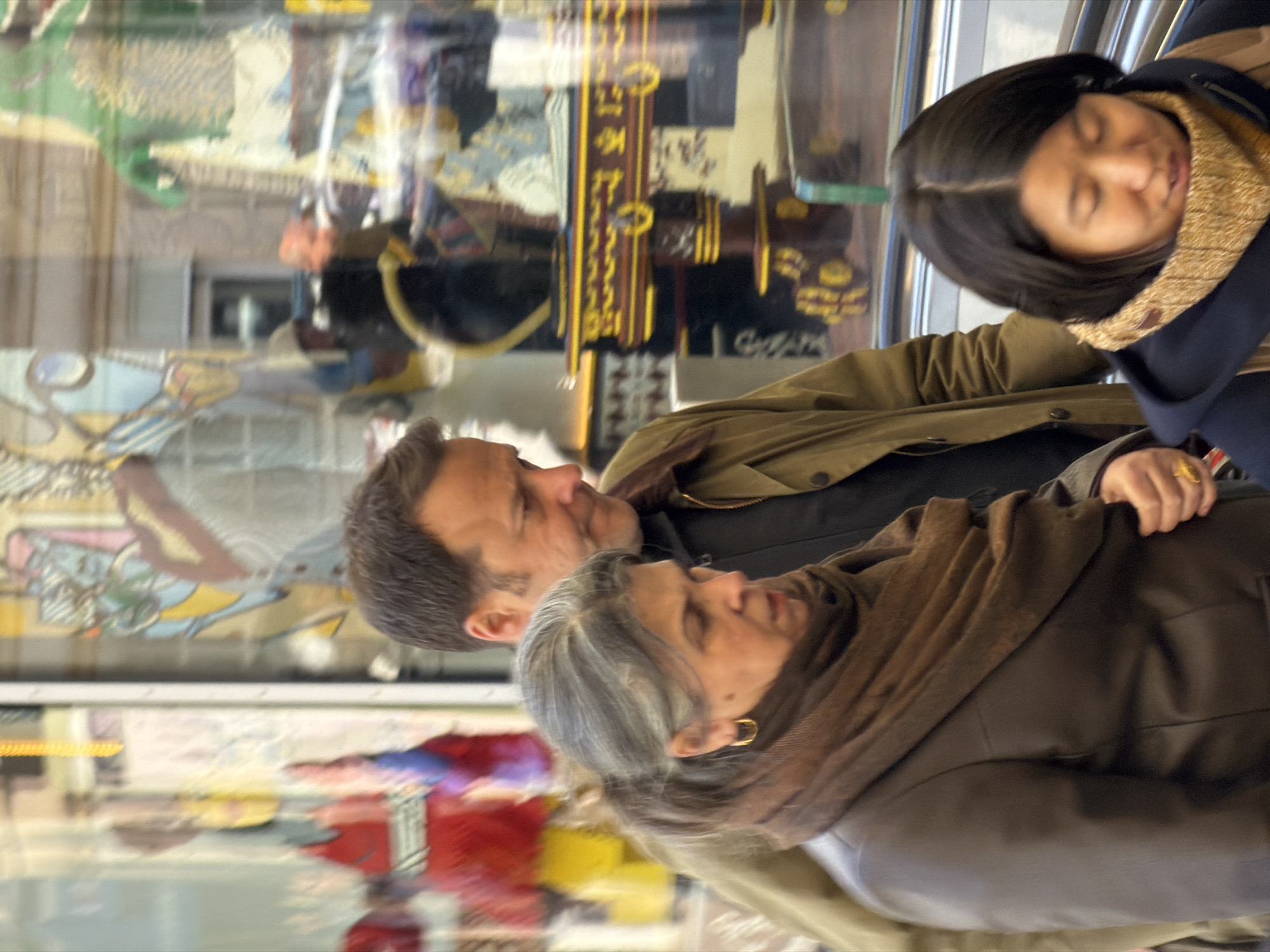 The image depicts three individuals in an indoor public space, likely a department store, in Paris, France. An escalator railing is visible in the lower right. In the foreground, a young East Asian woman with dark hair, wearing a navy blue coat and a golden-brown knitted scarf, looks downward. Slightly behind and to her left is an older woman with salt-and-pepper hair, wearing a dark brown jacket and a dark brown scarf, adorned with gold hoop earrings and a ring on her left ring finger; she looks towards her right. Behind the older woman and to her right stands a Caucasian man with short dark hair, dressed in an olive-brown jacket over a dark top, looking down towards the older woman. He wears a gold ring on his left ring finger. The background consists of highly reflective surfaces, displaying colorful and ornate artistic installations or decorative objects, indicating elaborate window displays of a commercial establishment. The scene suggests general movement, possibly on an escalator, with no direct interaction evident between the main subjects.