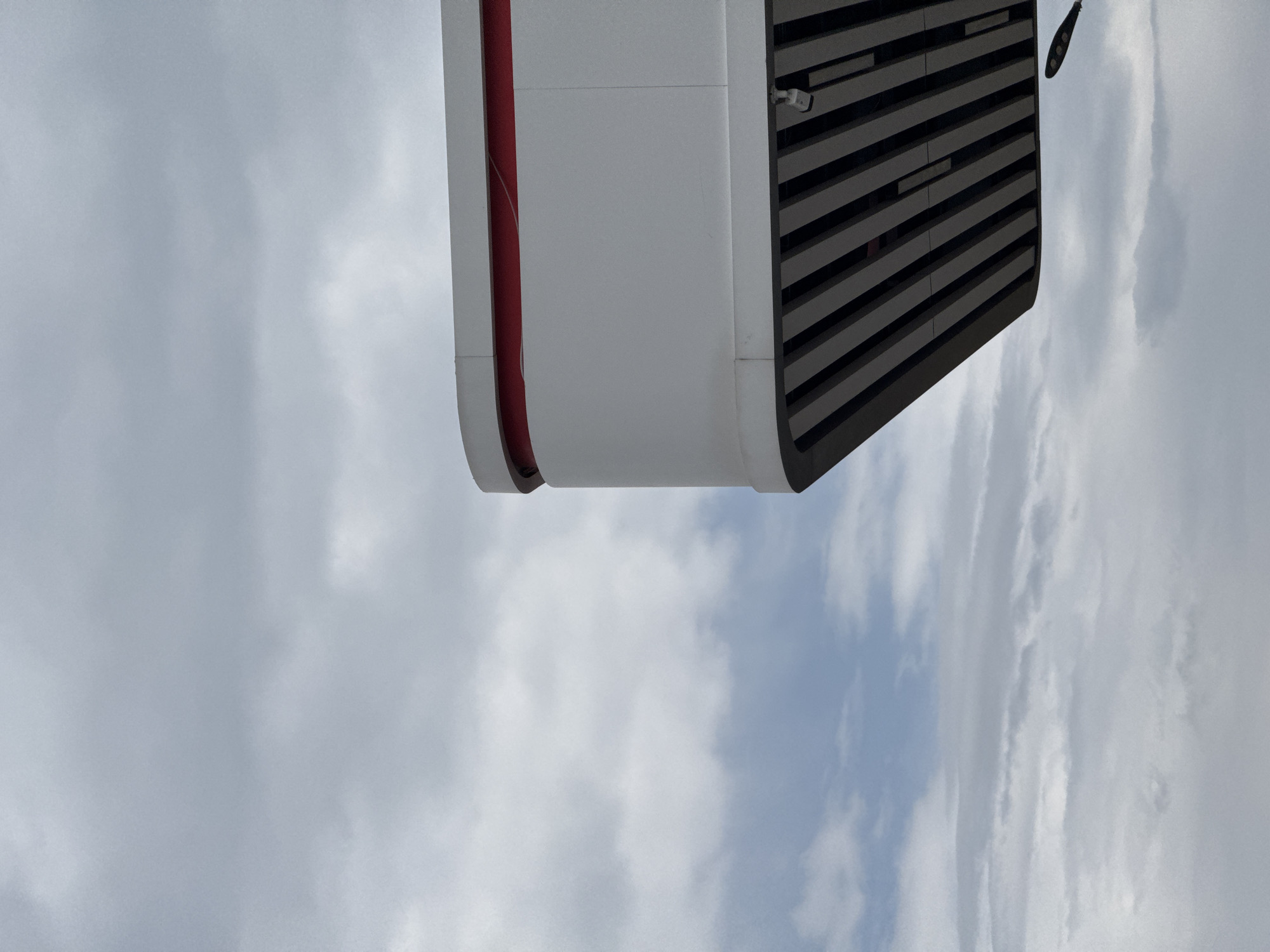 The image shows a close-up, low-angle shot of the canopy of a modern gas station in Balykchy, Kyrgyzstan. The canopy is white with a red accent stripe and features a slatted underside. A security camera is visible mounted on the underside. The sky is overcast with a mix of gray and white clouds. A streetlamp is also visible in the lower right corner of the frame. The overall impression is one of utilitarian modern architecture against a cloudy sky.