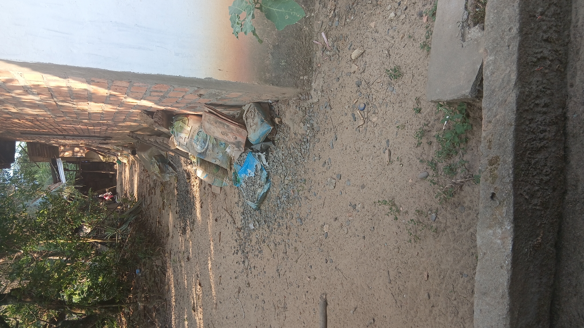 A dirt path runs alongside a brick building in Prabumulih, Indonesia. The path is uneven and covered in gravel and small stones. To the right of the path, a pile of building materials, including sacks of cement and a blue tarp, is leaning against the brick wall. The building itself appears to be under construction, with exposed brickwork and a partially completed white wall. Trees and vegetation are visible on the left side of the path, creating a dappled shade. In the distance, a person can be seen walking along the path. The scene appears to be an outdoor storage or construction area.