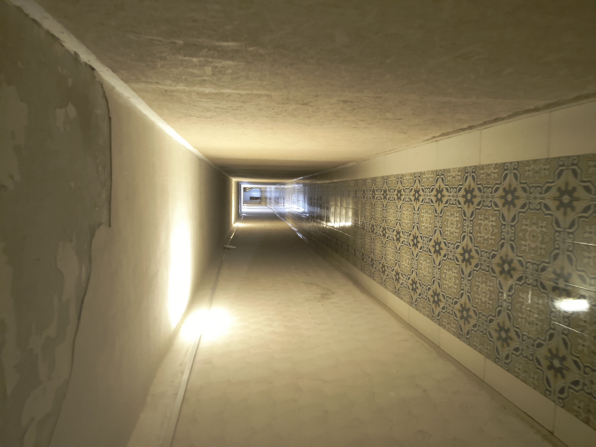The image depicts a long, dimly lit corridor with a patterned tiled floor. The walls are a neutral, textured color and appear to be made of plaster. The ceiling shows some signs of wear and tear, with visible patches and cracks. The corridor is illuminated by lights along the ceiling, creating reflections on the polished floor. The perspective suggests the photo was taken from one end of the corridor, looking towards the other end where a faint light and a silhouette of a person can be seen. The overall impression is of an interior passageway, potentially in a hotel or a similar establishment, in the city of Rivne, Ukraine.