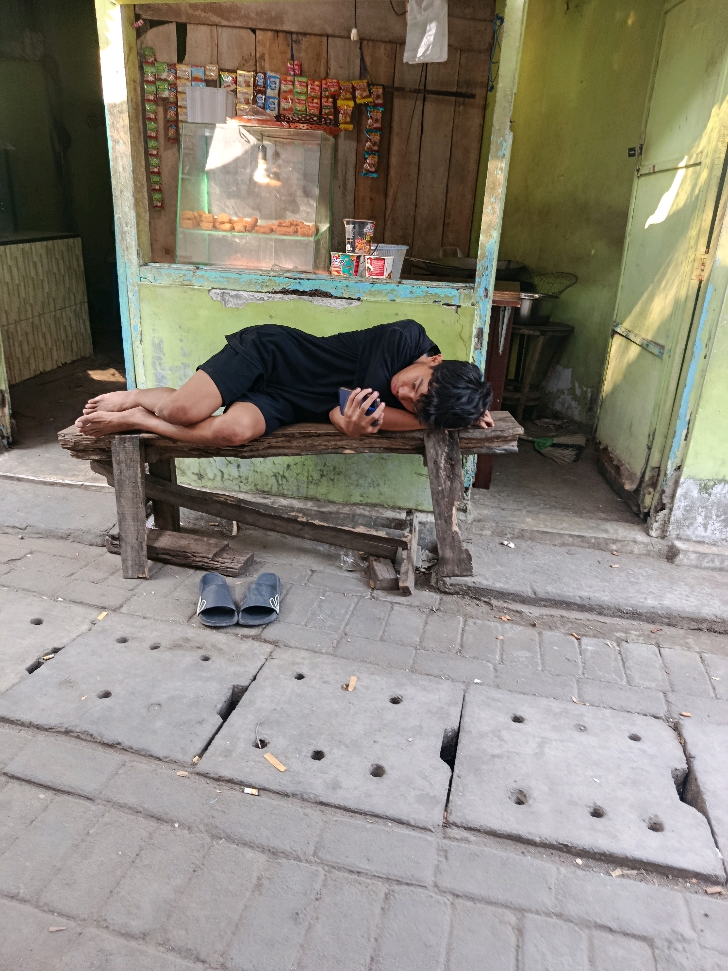 A young male, possibly a teenager, wearing a black t-shirt and black shorts, is sleeping on a rustic wooden bench in front of a small, open-air stall. The stall appears to be a roadside kiosk or warung, likely located in a residential area of Jobokuto, Indonesia, given the style of construction and the presence of concrete slabs with drainage openings in the foreground, typical of Indonesian urban infrastructure. The stall has a counter with a glass display case showing what appear to be fried snacks or pastries. Various packaged goods are displayed on shelves and hanging from the wooden structure behind the counter. The stall's walls are painted a light green, and the wooden elements are weathered. A pair of dark-colored flip-flops are placed on the ground in front of the bench. The scene is outdoors, with sunlight casting shadows. The ground is paved with interlocking grey bricks and concrete slabs. The overall impression is of a simple, everyday scene in a working-class urban environment.