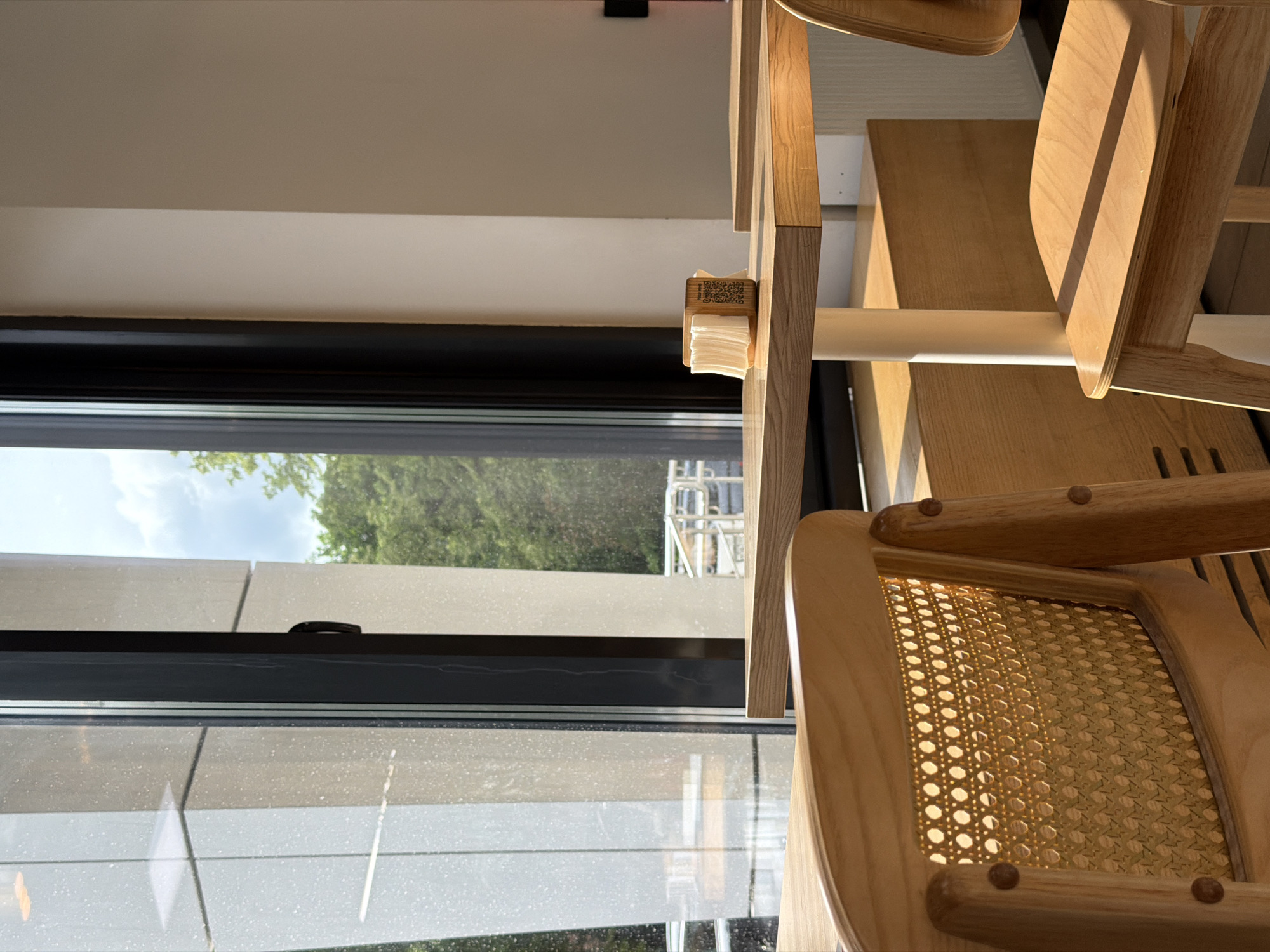 The image captures an interior scene where light wooden furniture is positioned near a large window. Through the window, one can see trees, a cloudy sky, and a partial view of a structure resembling scaffolding. A small dispenser containing tissues is attached to the table. The location in Izhevsk, Russia, cannot be determined from the image. There are no people visible.
