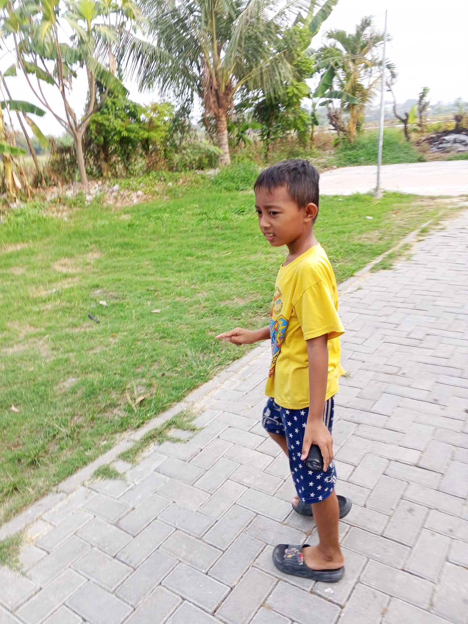 A young boy wearing a yellow t-shirt and blue shorts with white stars walks on a paved pathway. He is holding a black object in his right hand. The pathway is bordered by a grassy area with trees and foliage in the background. The scene appears to be outdoors, possibly in a park or residential area. The setting is consistent with a location in Ciruas, Indonesia, given the tropical vegetation.