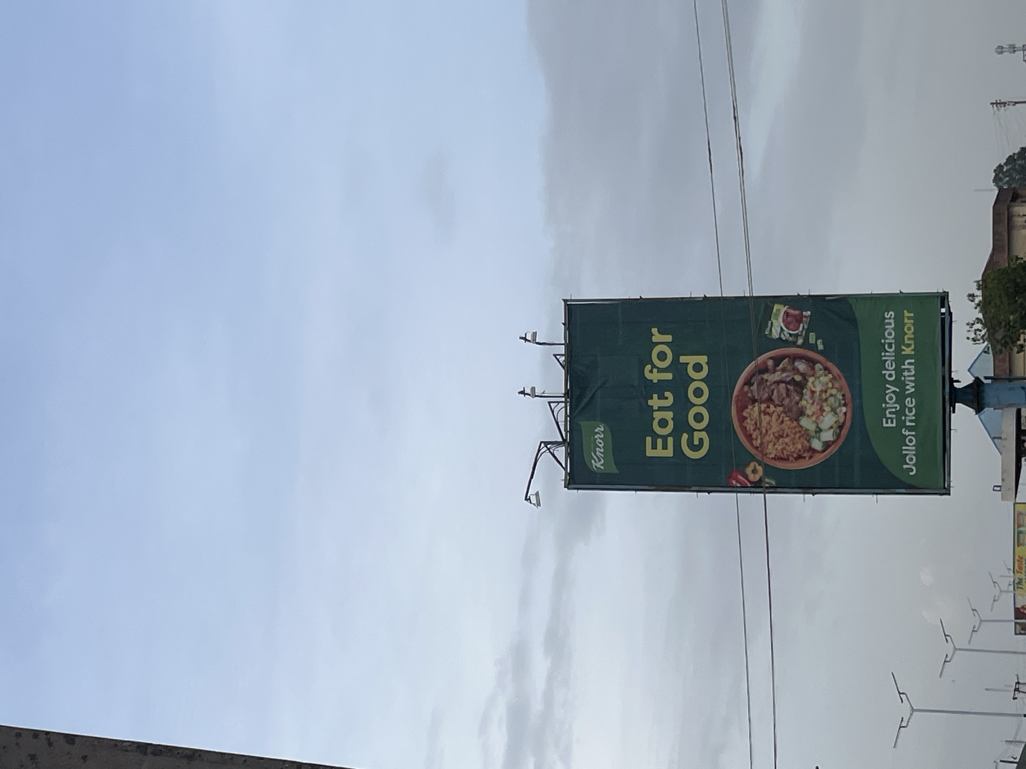 The image shows a billboard for Knorr, a food brand. The billboard is green and features a bowl of jollof rice, a popular West African dish. The text on the billboard reads, "Eat for Good" and "Enjoy delicious Jollof rice with Knorr." There is also a small Knorr logo on the billboard. The billboard is supported by a metal structure and is positioned outdoors, likely along a road. The sky in the background is overcast, suggesting it might be daytime, possibly in the morning or late afternoon. There are power lines visible above the billboard. In the background, there are some buildings and trees, indicating an urban or semi-urban setting. No people are clearly visible in the image, but the overall scene suggests a commercial advertisement in a public space. The weather appears to be cloudy.