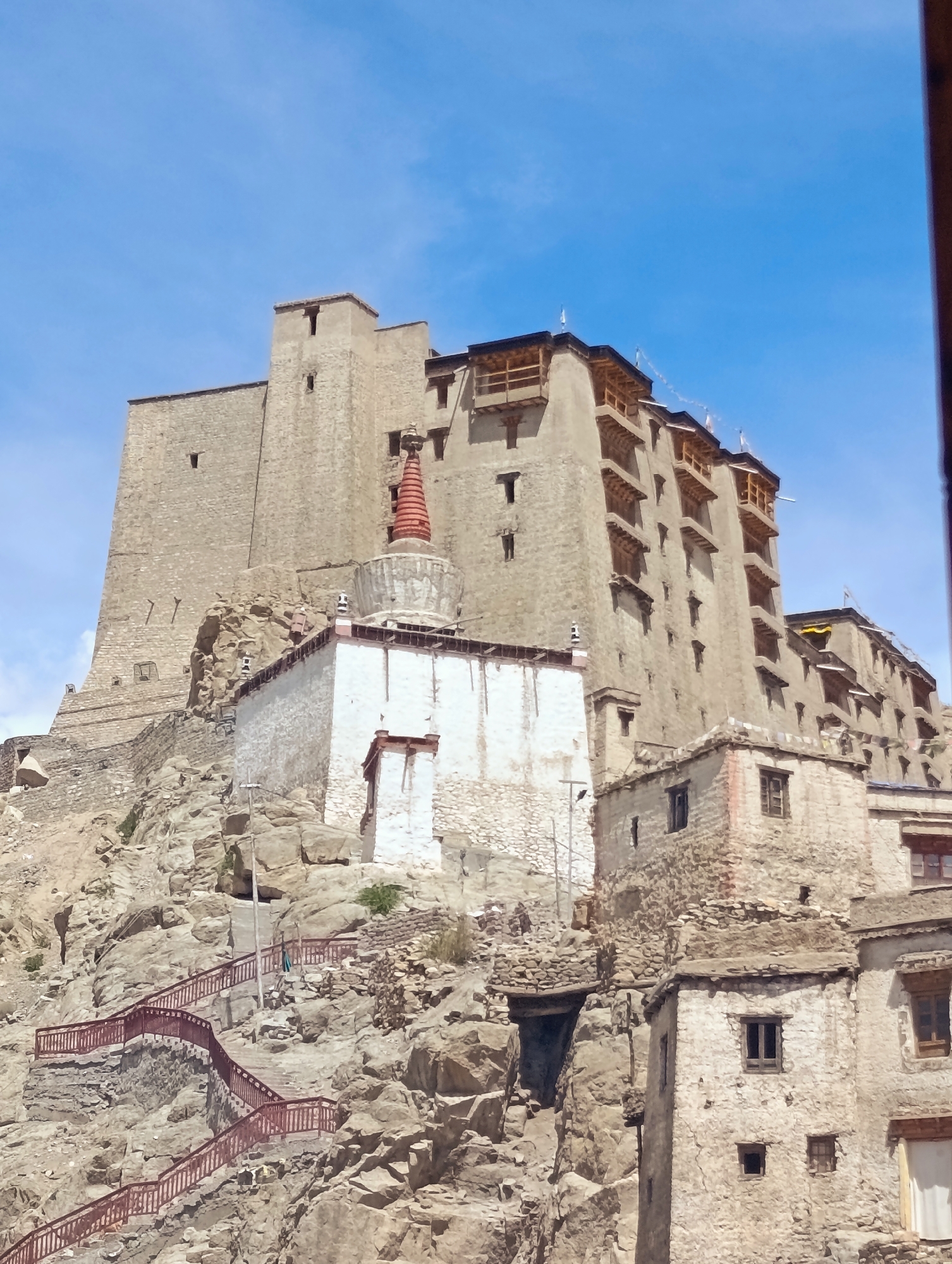 The image shows the Leh Palace, a nine-story royal palace and monastery complex in Leh, Ladakh, India. The palace is situated on a rocky hill overlooking the town of Leh. The structure is made of stone and wood and features intricate carvings and paintings. In the foreground, a set of red-railed stairs winds up the steep rocky slope, leading towards the palace. The sky is a clear blue with a few wispy clouds. The architecture of the palace is a blend of Tibetan and Kashmiri styles, with its whitewashed walls and multi-tiered roofs. The image captures the grandeur and historical significance of this iconic landmark.