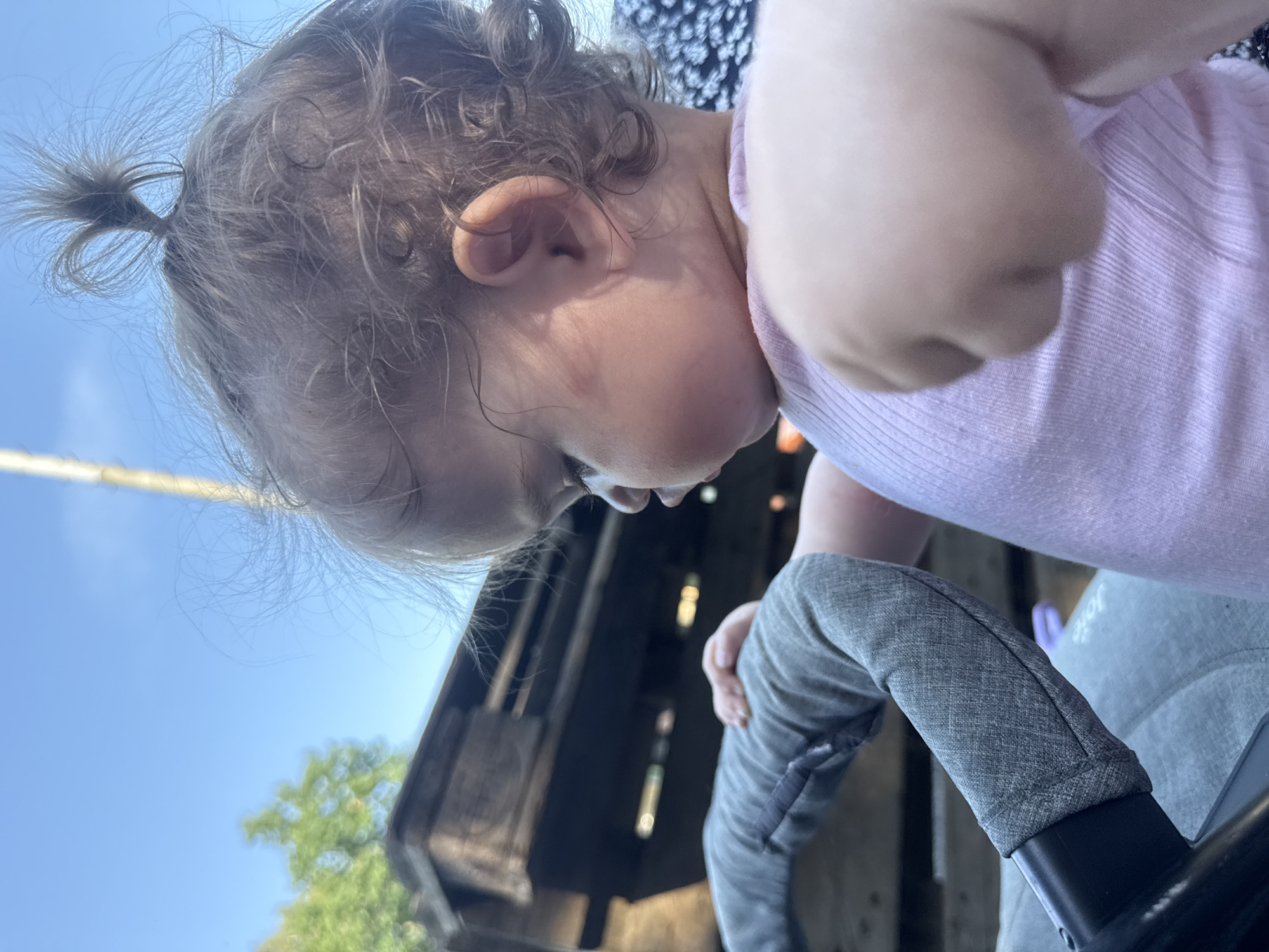 A close-up, eye-level outdoor shot shows a toddler, viewed from the right side, looking down and to the left. The toddler has light brown curly hair styled in a small ponytail on top of their head, and is wearing a pink ribbed tank top. Their left arm is visible, bent at the elbow, with their fist clenched. The toddler is seated in a stroller, with a gray padded handle visible in the foreground. In the background, out of focus, is a wooden structure and a clear blue sky. The scene is set in Beltershain, Germany.