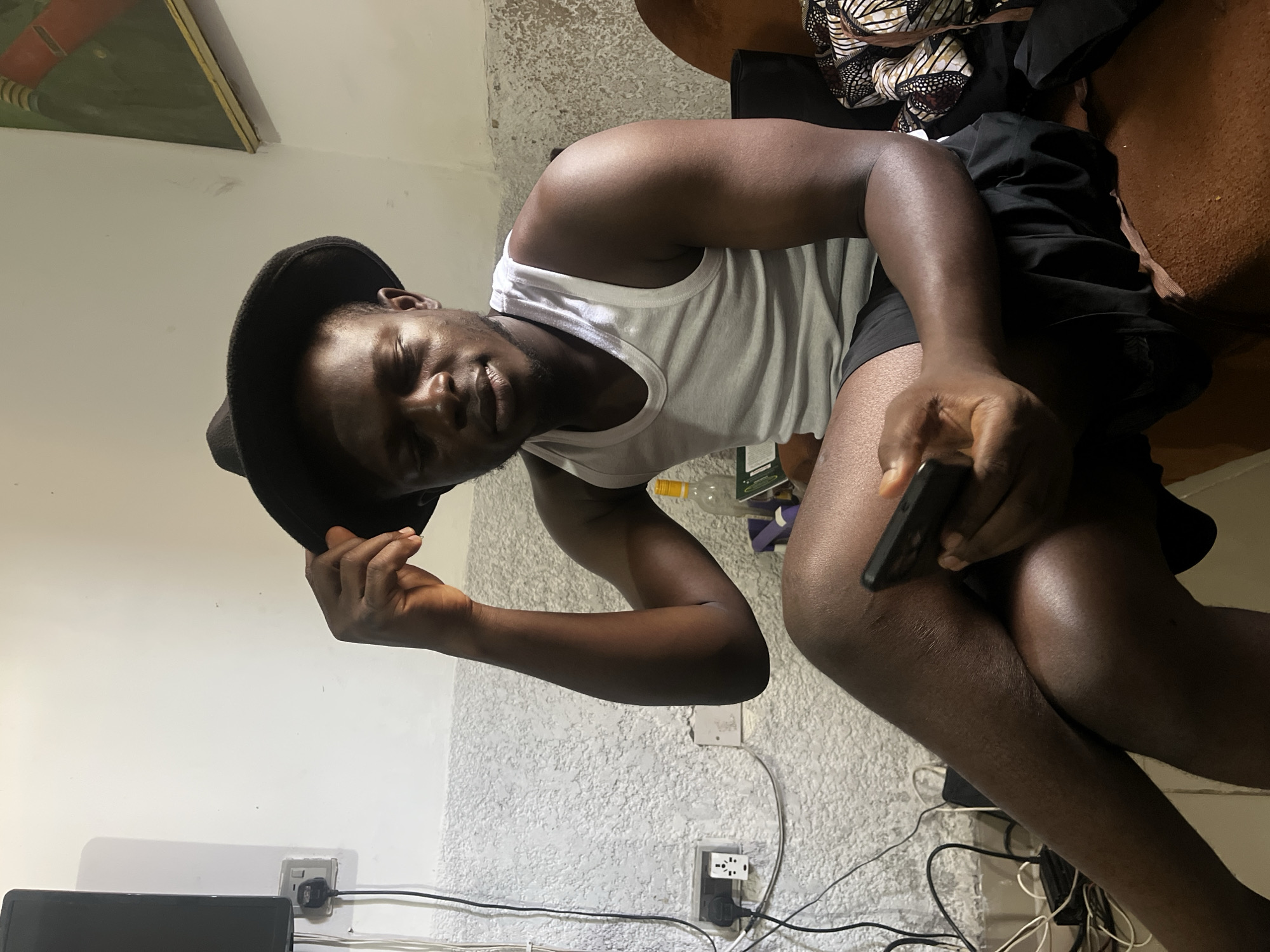 An indoor scene in Abuja, Nigeria, depicts a dark-skinned male seated on a worn, brown upholstered couch. He wears a black fedora-style hat, a white tank top (singlet), and dark shorts. His right hand adjusts the brim of his hat, while his left hand holds a black smartphone, which he gazes down at. His legs are crossed at the knee. The background features light-colored walls; the lower portion exhibits a textured, off-white finish, transitioning to a smooth white upper section. On the left wall, a black flat-screen television or monitor is partially visible above multiple electrical outlets with attached plugs and loose black and white wiring. To the right, a portion of a framed artwork featuring red and green elements is mounted on the wall. Behind the man's right shoulder, a clear bottle containing a yellowish liquid, a green object, and other indistinct items are present on a surface. A pile of patterned fabric and black clothing items rests on the couch beside him.