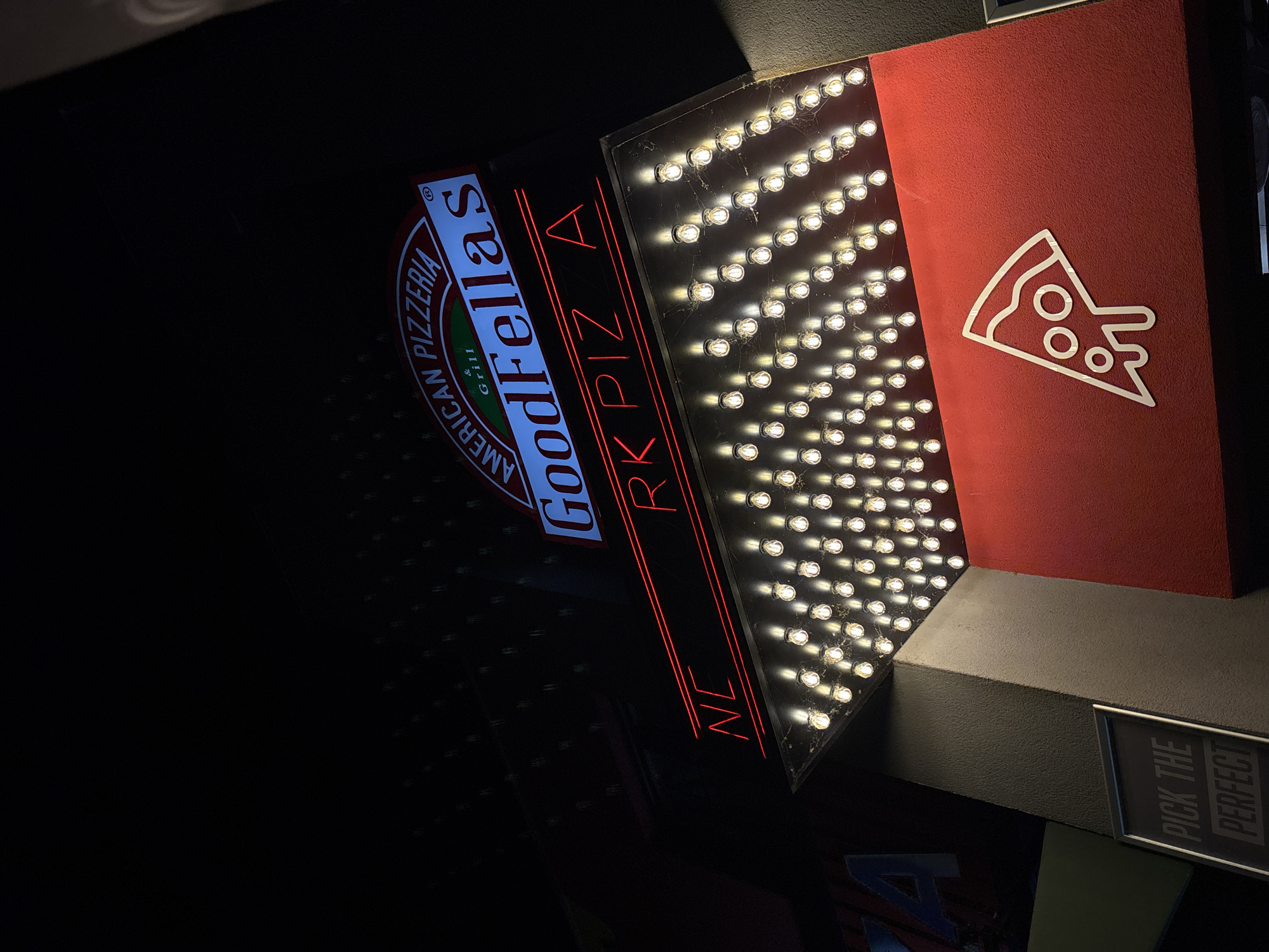 Illuminated signs for the Goodfellas pizzeria in Podgorica, Montenegro. The sign includes: the words "Goodfellas American Pizzeria & Grill"; the neon letters "NCRK PIZZA" in red; a light board with many small lights; and a red board with a white pizza slice. A sign reading "Pick the Perfect" is visible.