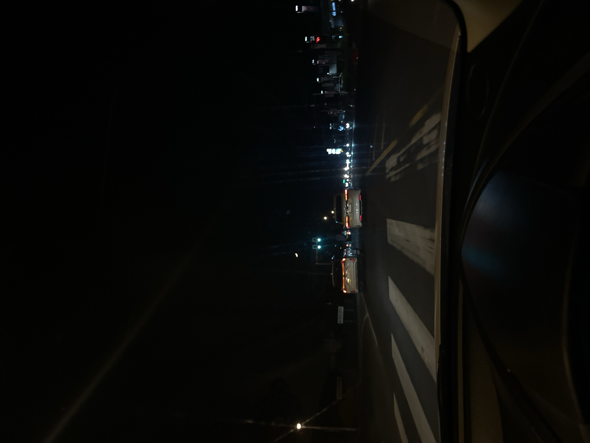 This is a nighttime, low-angle shot taken from inside a vehicle, likely looking out the windshield or side window. The image is dominated by darkness, with artificial lights providing the only illumination. The perspective is tilted, suggesting the camera may have been held at an unusual angle or that the vehicle itself is on an incline.
In the distance, a row of buildings with illuminated windows and signs can be seen, hinting at an urban or suburban environment. Several cars are visible on a road, their headlights and taillights creating streaks of light in the dark. One white vehicle, possibly an SUV or van, is particularly clear, with its red taillights glowing. Further down the road, traffic lights show green signals.
The foreground on the right is occupied by the interior of the vehicle, which appears to be a dark, curved surface, possibly part of the dashboard or interior trim. Some diagonal stripes of light, likely from streetlights or building lights, are reflected or cast onto this surface, creating a dramatic play of light and shadow. The overall scene evokes a sense of driving at night through a lit area.
There is no visible text in the image. The time of day is clearly night, and the weather is not discernible. The location context is Wichit, Thailand.