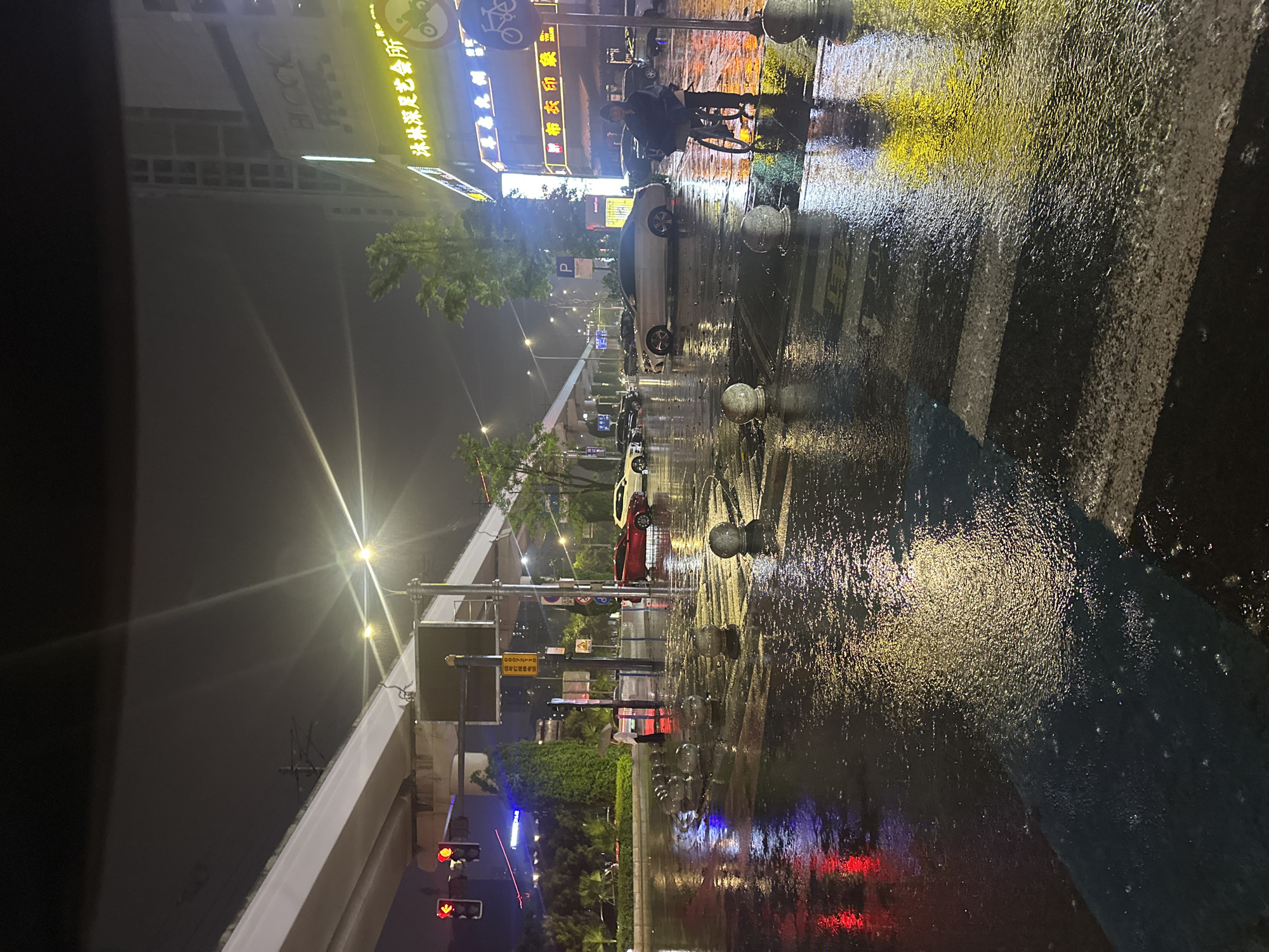 The image depicts a wet street scene in Chongqing High-tech Zone, China, at night. Rain has created a reflective surface on the pavement, mirroring the lights from buildings and streetlights. A person on a bicycle is visible near a pedestrian crossing. Several parked cars line the street, and a traffic light displays a red signal. Buildings with illuminated signs are visible in the background, written in Chinese. The presence of trees suggests a mix of urban and natural elements in the environment. A pedestrian crossing is marked on the road.