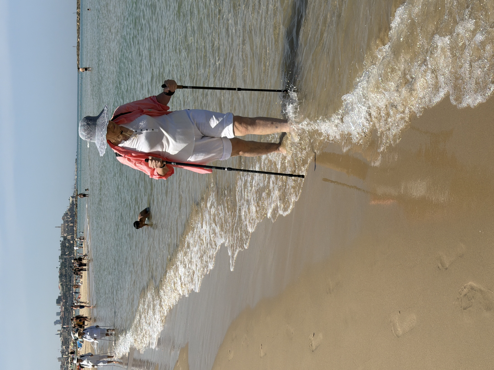 The scene is a sunny beach in Tel-Aviv, Israel. An elderly woman wearing a light blue patterned hat, white shirt and shorts, and a coral-colored jacket is wading into the water with the aid of two black walking sticks. The water is clear and the waves are gentle, washing onto the tan-colored sand. Several people are visible in the background along the beach, some in the water and some on the sand. The Tel-Aviv skyline is visible in the distance. Footprints dot the sand in the foreground.