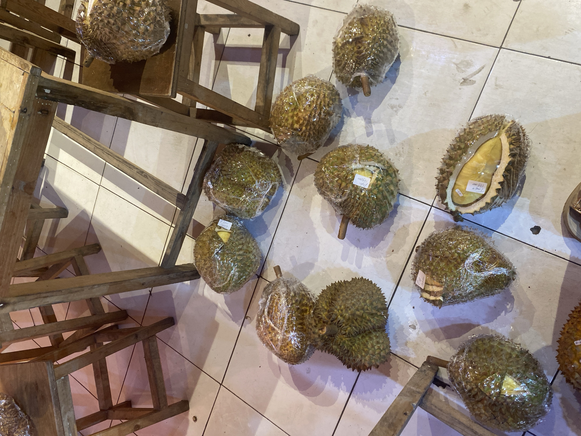 The image depicts a close-up, high-angle view of several durians, a spiky tropical fruit, displayed on a tiled floor in what appears to be an outdoor market or food stall. The durians are individually wrapped in clear plastic. Some of the fruits are whole, while one in the foreground has been cut open, revealing its yellow flesh. Several wooden stools and table legs are visible in the background, suggesting a seating or serving area. The lighting is somewhat dim, creating shadows on the floor. The overall impression is of a retail setting where durians are being sold. The scene is likely in Depok, Indonesia, given the common availability and popularity of durians in that region.