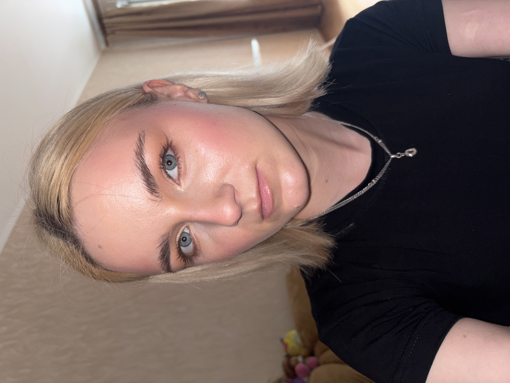 A young woman with blonde hair and blue eyes is taking a selfie indoors. She is wearing a black t-shirt and a silver necklace. Her makeup is visible, including eyeshadow and blush. The background shows a wall with a textured pattern and a window with curtains. There is also a hint of furniture and stuffed animals in the background. The image appears to be taken in a residential setting in the city of Sinyavino, Russia.