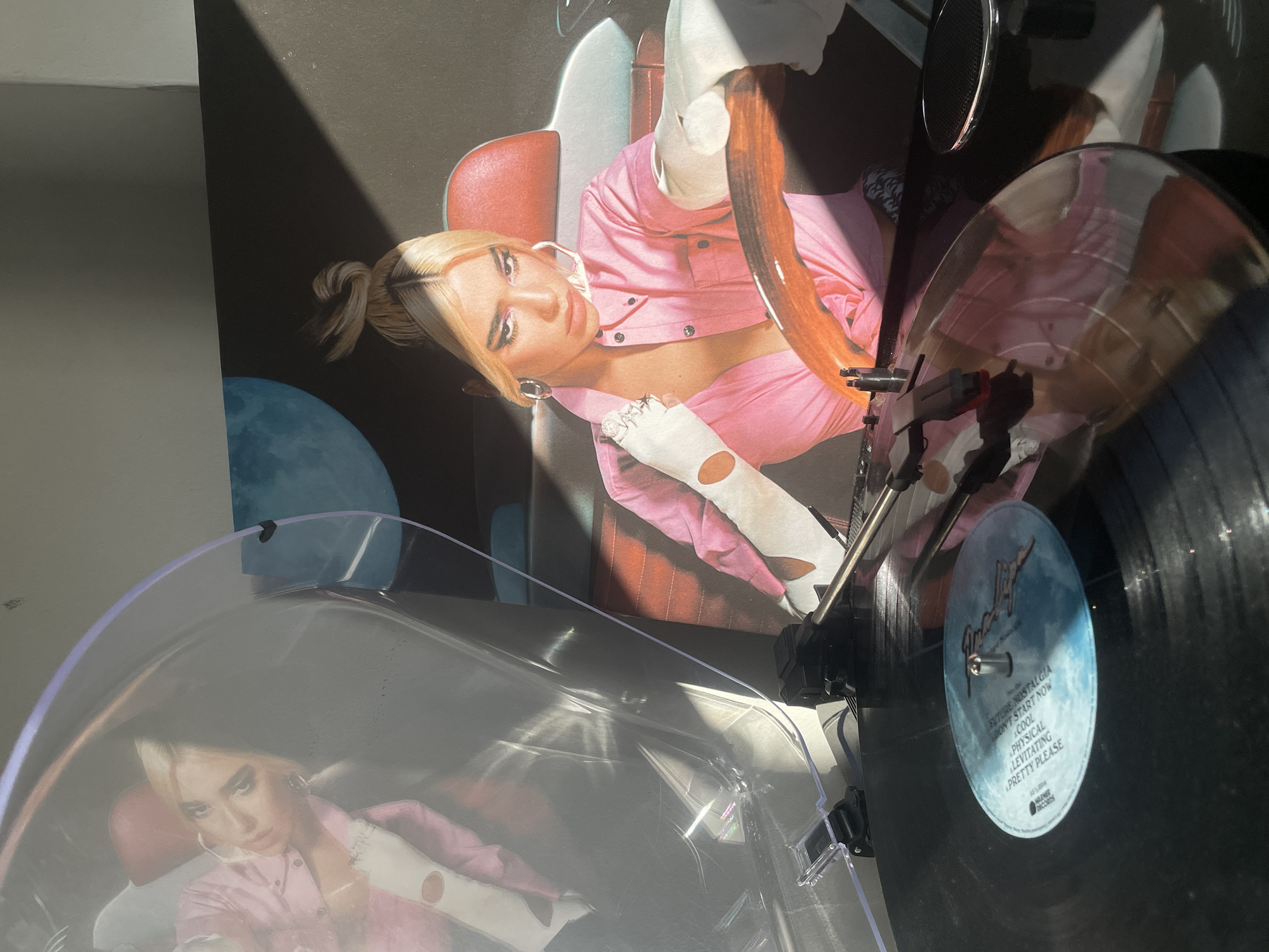 The image shows a vinyl record player playing a vinyl record. The album cover for Dua Lipa's "Future Nostalgia" is placed behind the record player. The record is black with a blue label. The room appears to be indoors and well-lit.