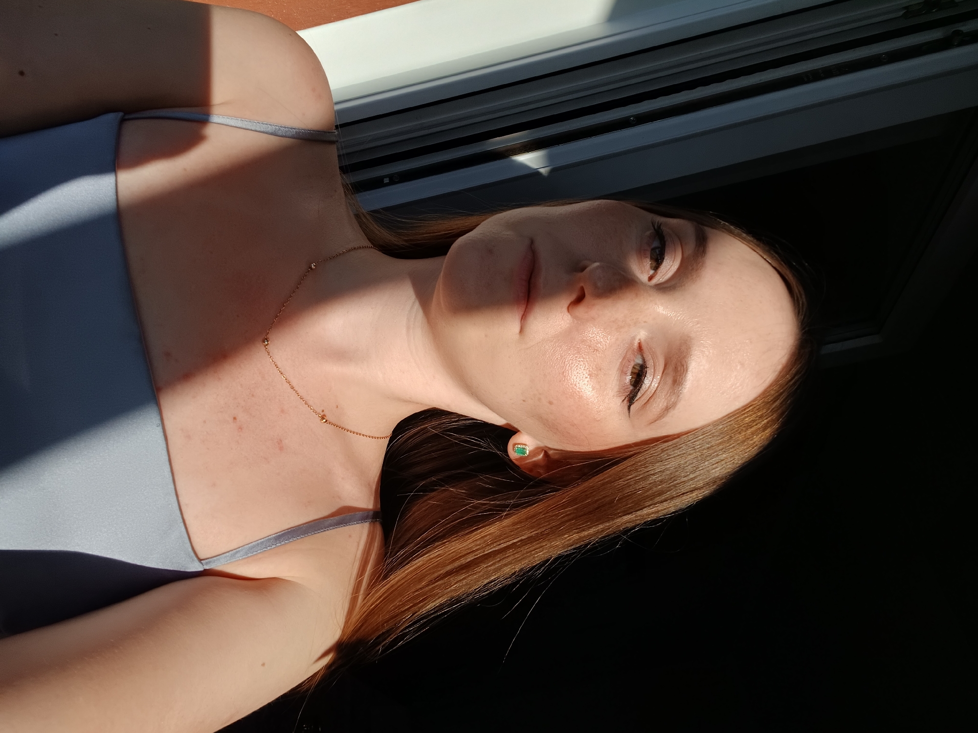 This is a close-up, rotated media file of a young person's upper chest and face. The person has fair skin with freckles, light brown hair, and is wearing a light blue camisole. A thin gold necklace with small beads is visible around their neck. They are also wearing a small, rectangular green stud earring.
The scene is lit by natural sunlight, casting a strong shadow across the person's chest and shoulder. The lighting suggests it is either early morning or late afternoon. The background is divided into two parts: a lighter area with horizontal blinds, possibly a window, and a darker area which is out of focus. The overall mood appears relaxed and casual. No text is visible in the media file.